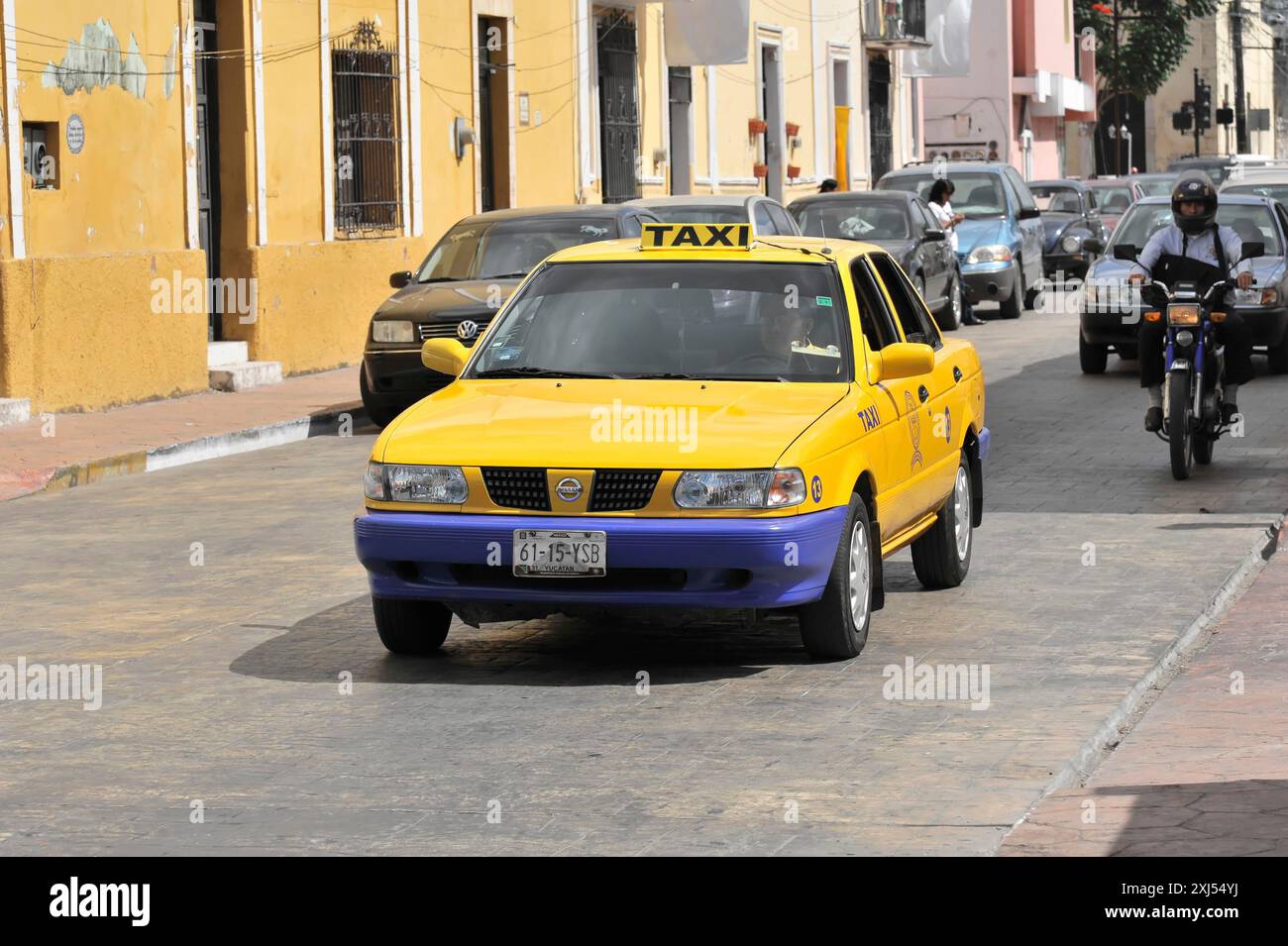 Merida, Yucatan, Mexiko, Mittelamerika, gelbes und blaues Taxi in einer belebten Stadtstraße mit Fahrzeugen und Gebäuden Stockfoto