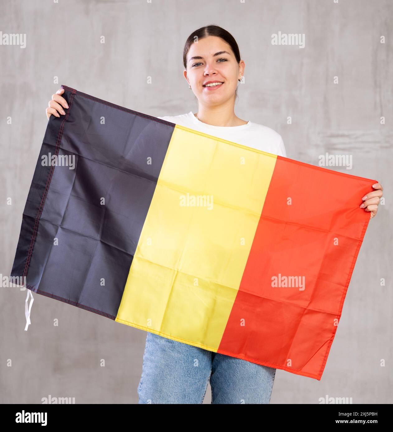 Fröhliche junge Frau, die posiert und lächelt, mit der belgischen Flagge an der grauen Wand steht Stockfoto