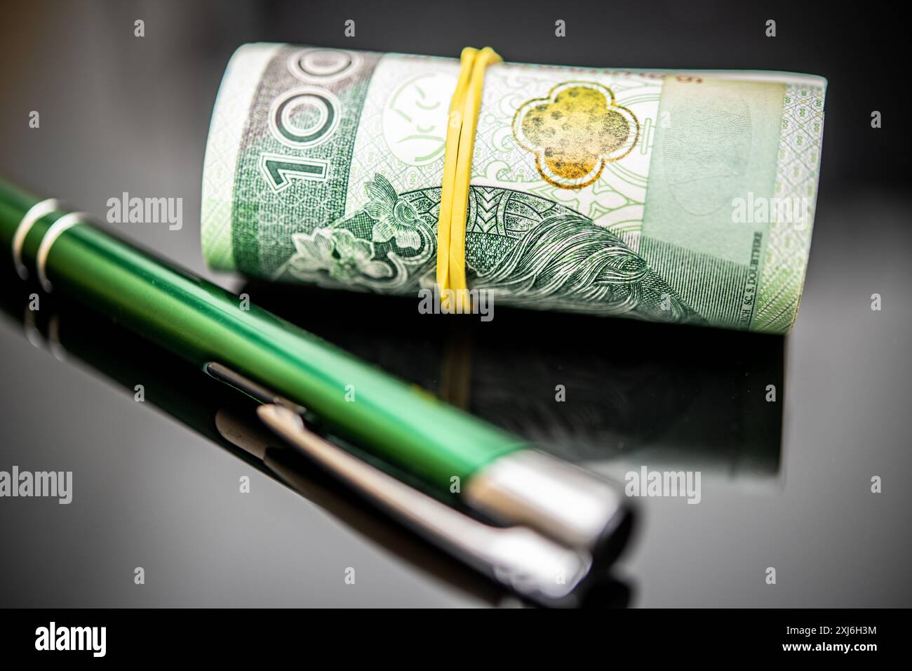 Polnische Währung, 100 Zloty-Banknoten gerollt und gesichert mit einem gelben Gummiband, neben einem eleganten grünen Stift auf glänzendem Hintergrund, emphasi Stockfoto