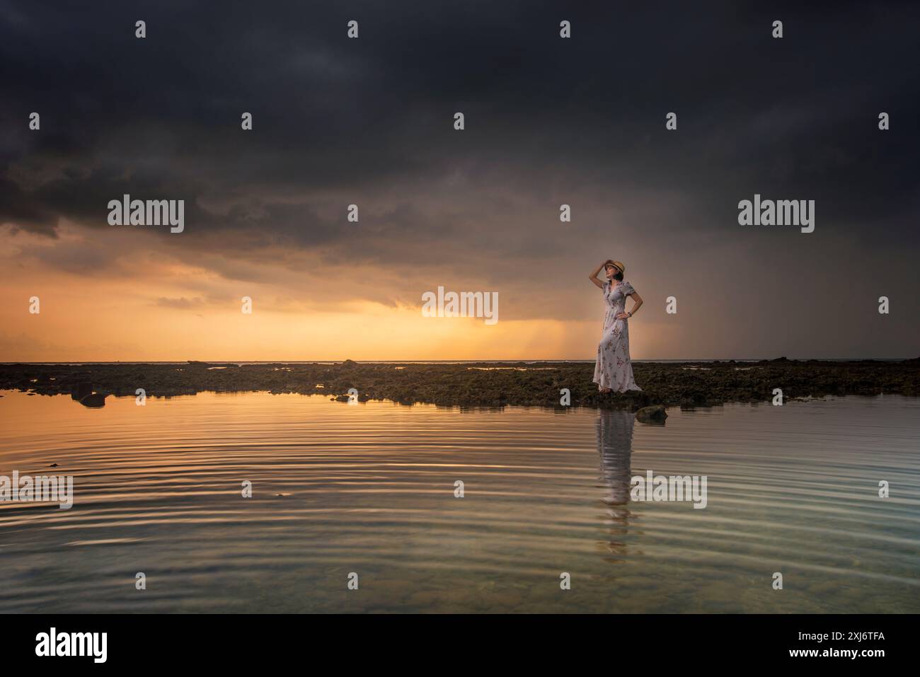 Lächelnde Frau in einem langen Sommerkleid, die bei Sonnenuntergang an einem felsigen Strand steht, Indonesien Stockfoto