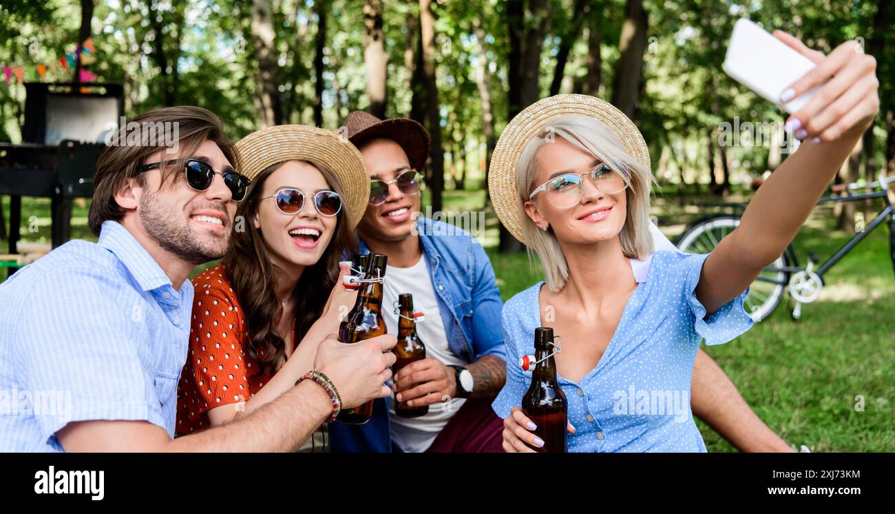 Lächelnd multikulturellen Freunde mit Bier unter selfie auf dem Smartphone während im Sommer Park ruhenden Stockfoto