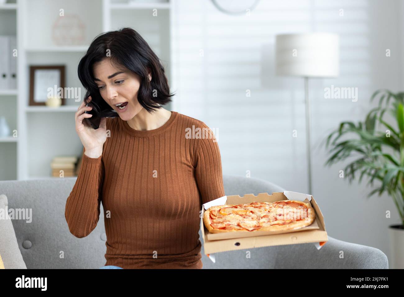 Wütende junge Frau, die zu Hause steht, eine Pizzaschachtel hält und wütend am Telefon spricht und sich über den Lieferservice beschwert. Stockfoto