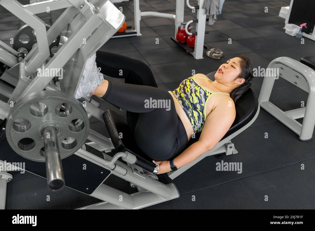 Junge übergewichtige Frau, die in einem örtlichen Fitnessstudio mit einer Beinpresse an ihren Beinen arbeitet Stockfoto