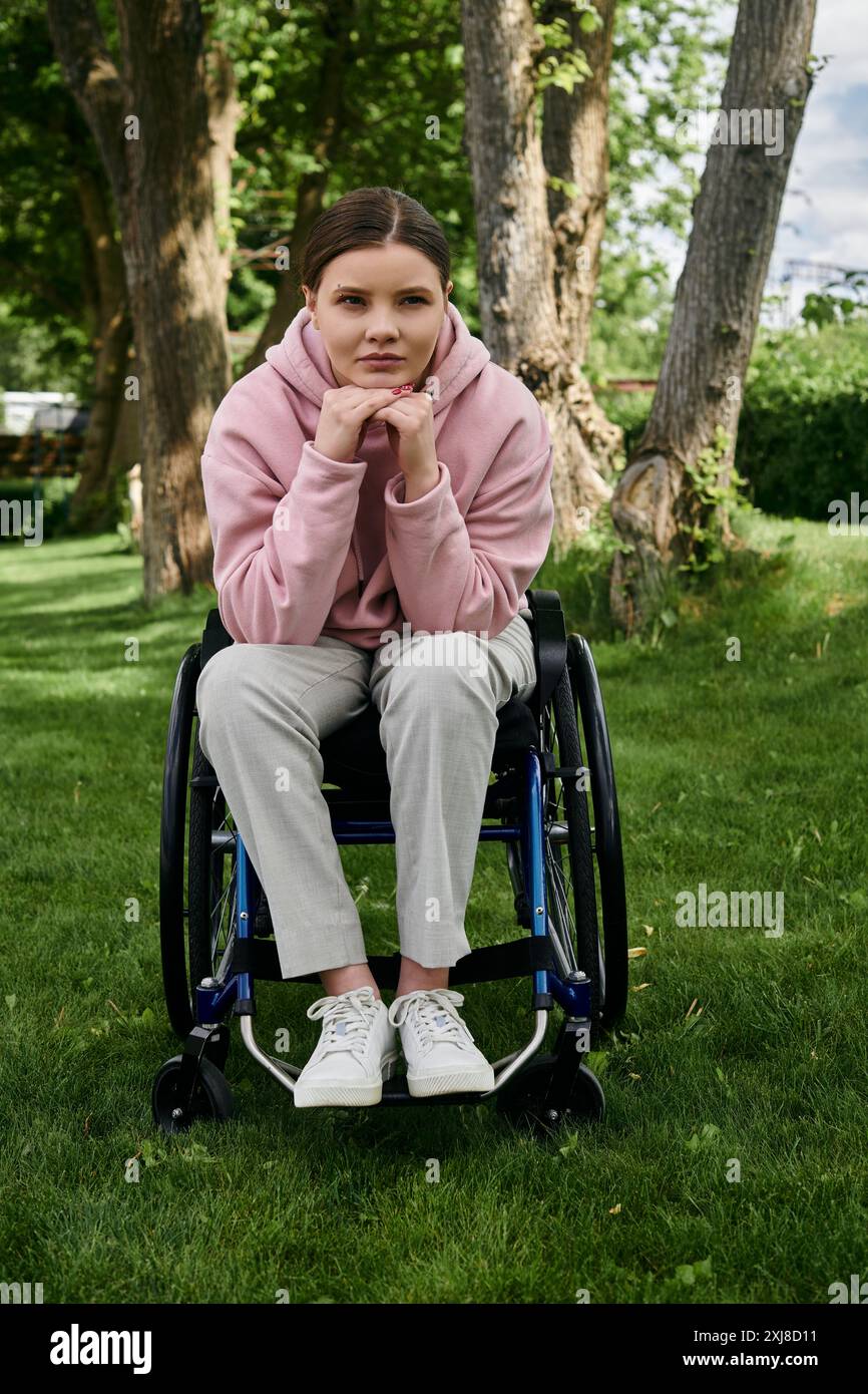 Eine junge Frau in einem rosa Hoodie sitzt in einem Rollstuhl in einem grasbewachsenen Park. Stockfoto