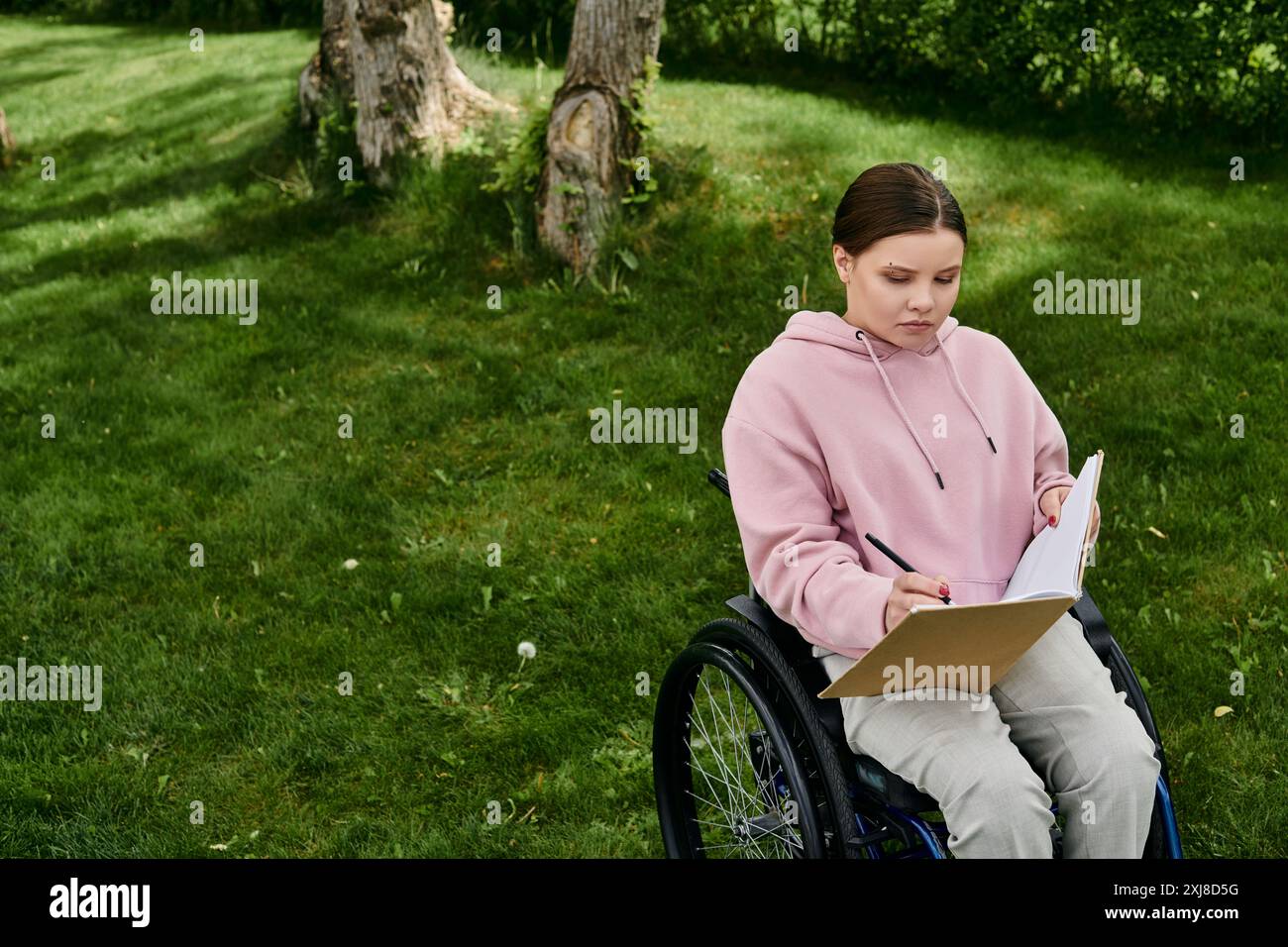 Eine junge Frau in einem rosafarbenen Hoodie sitzt im Rollstuhl und schreibt in ein Notizbuch, während sie einen sonnigen Tag im Park genießt. Stockfoto