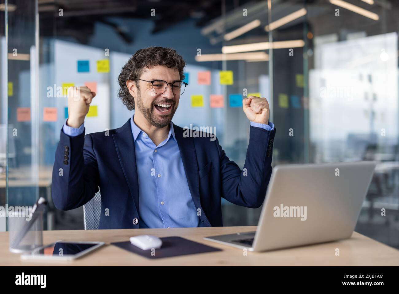 Geschäftsmann im Anzug feiert Erfolg am Schreibtisch mit Laptop und zeigt Aufregung und Freude. Professionelle Umgebung mit moderner Einrichtung, Highlight Stockfoto