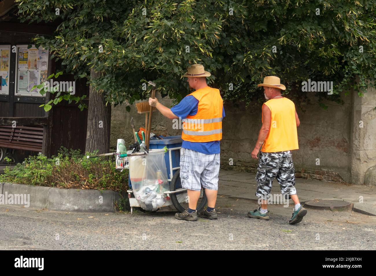 Zwei Straßenputzer in orangefarbenen Westen und Hüten arbeiten zusammen mit einem Straßenputzwagen auf einer Vorstadtstraße Stockfoto