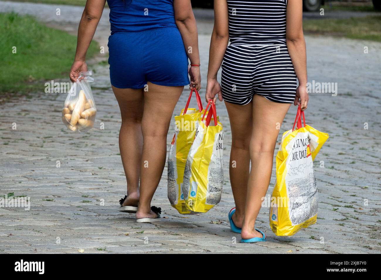Zwei Frauen gehen auf einem Kopfsteinpflasterweg mit gelben Einkaufstaschen und einer kleinen Plastiktüte mit Rollen Stockfoto