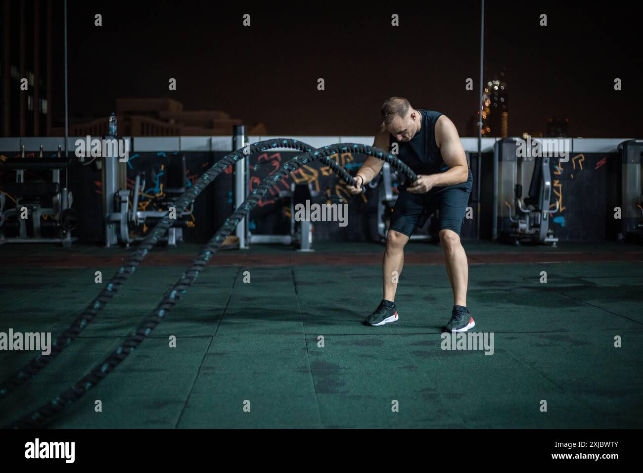 Ein Mann in einem schwarzen Tanktop und Shorts trainiert nachts in einem Fitnessstudio auf dem Dach mit Kampfseilen. Stockfoto