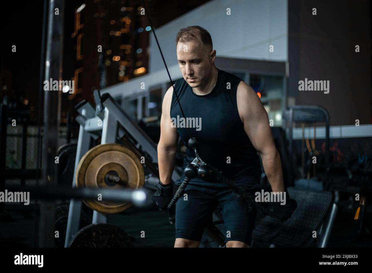 Ein Mann in einem schwarzen Tanktop arbeitet nachts an einer Kabelmaschine in einem Fitnessstudio. Stockfoto