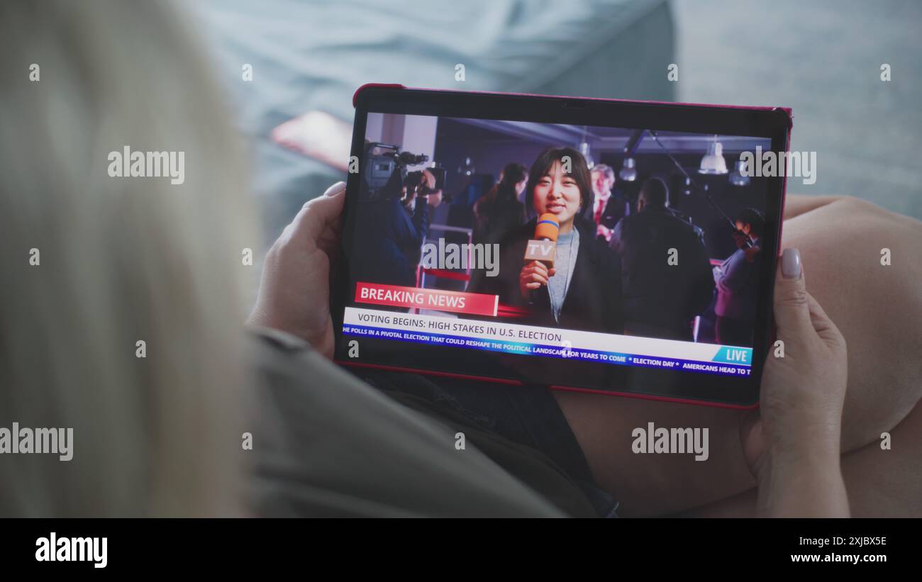 Tablet-Bildschirm zeigt die Wiedergabe von Nachrichten mit asiatischen Korrespondenten. Die kaukasische Frau sieht Nachrichtenberichte über US-Wahlen mit einem Tablet-Computer, während sie auf dem Sofa liegt. Nahaufnahme. Stockfoto