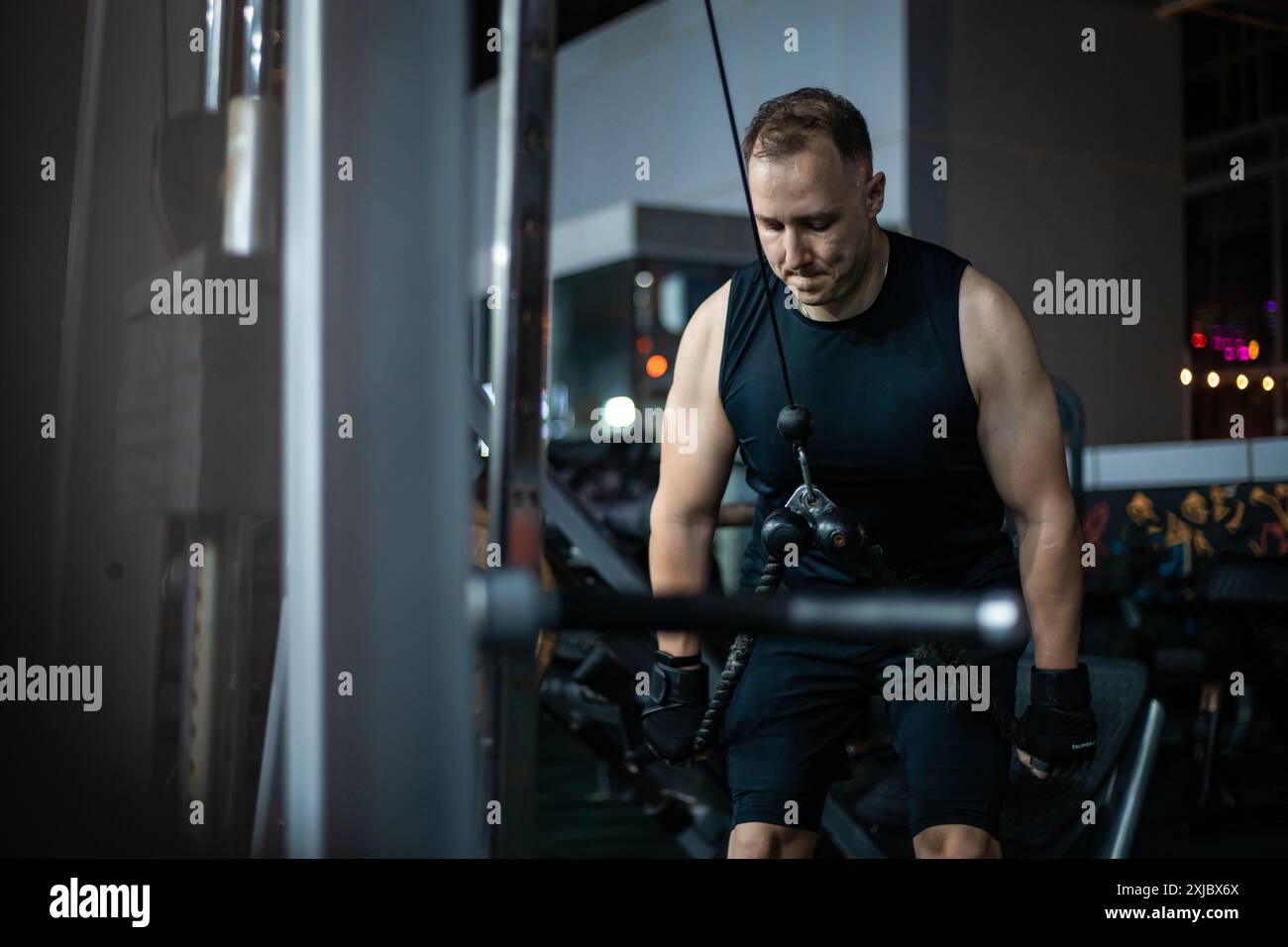 Ein Mann in einem schwarzen Tanktop und Shorts benutzt eine Kabelmaschine in einem Fitnessstudio. Stockfoto