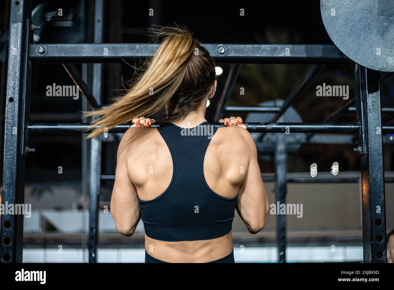 Eine Frau in einem schwarzen Sport-BH macht in einem Fitnessstudio Pull-Up-Bar. Ihr blondes Haar ist in einem Pferdeschwanz zurückgezogen. Stockfoto