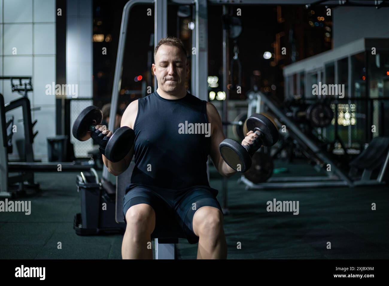 Ein Mann hebt nachts in einem Fitnessstudio Gewichte. Er trägt ein schwarzes Tanktop und Shorts. Stockfoto