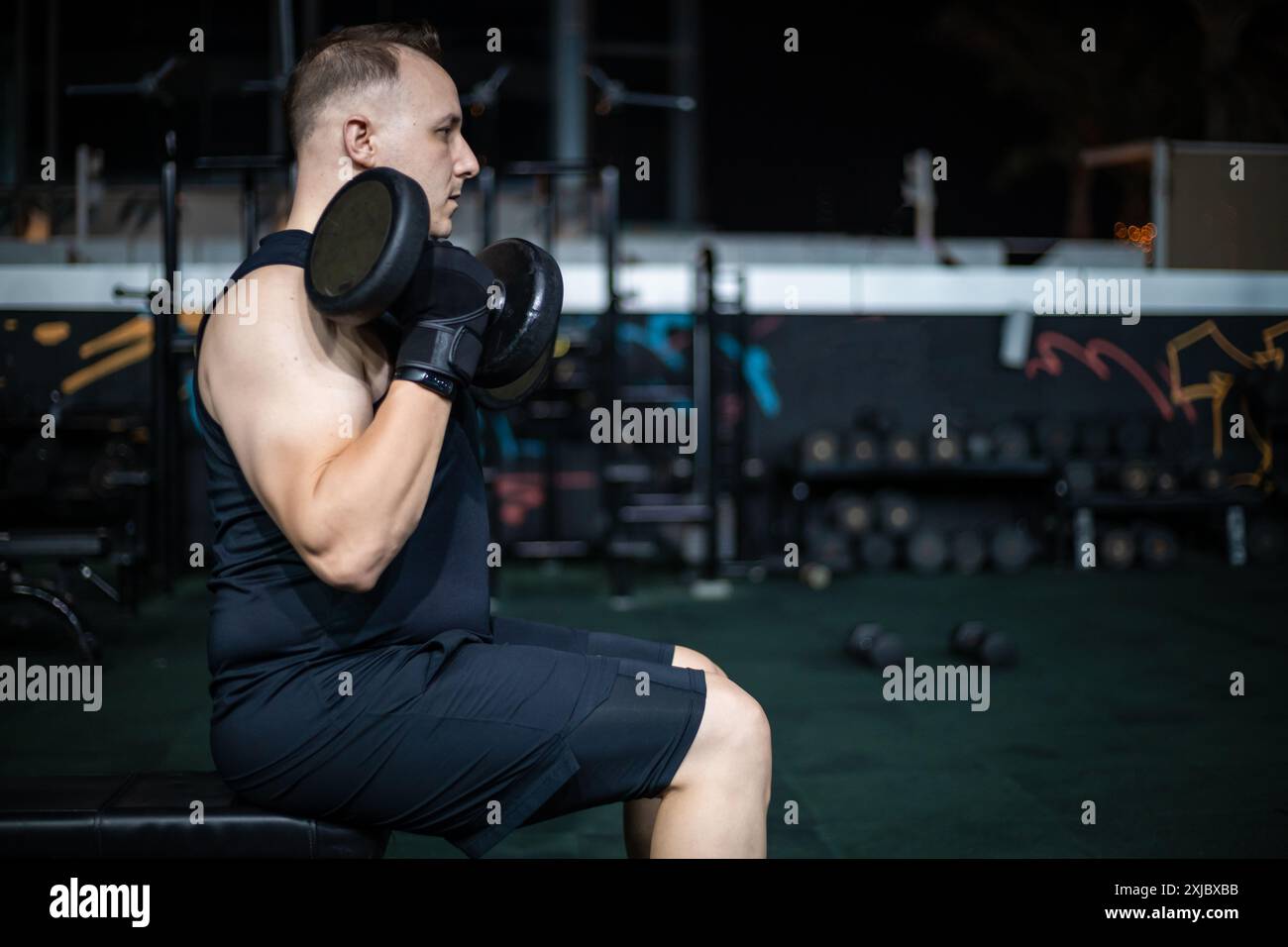 Ein Mann mit Tanktop und Shorts hebt Kurzhanteln, während er auf einer Bank im Fitnessstudio sitzt. Stockfoto