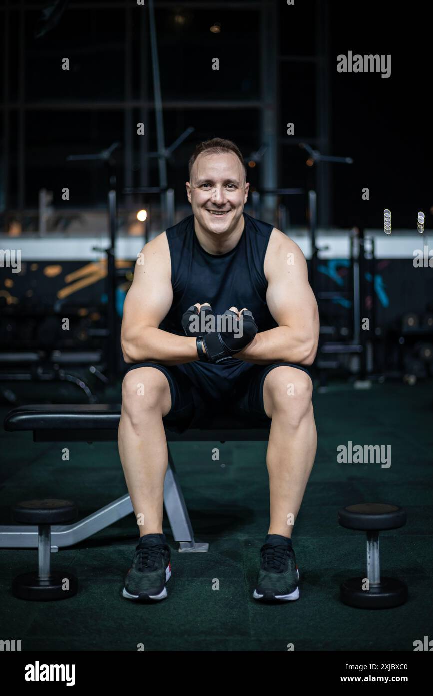 Ein Mann sitzt auf einer Bank in einem Fitnessstudio, trägt schwarze Trainingskleidung und hält schwarze Gewichte. Stockfoto