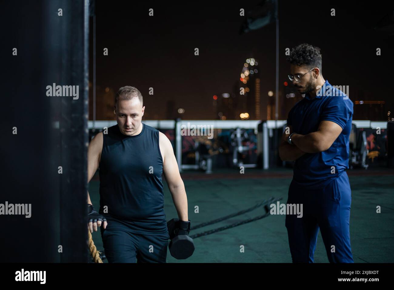 Ein Mann, der ein schwarzes Tanktop und eine schwarze Shorts trägt, hebt Gewichte, während ein Mann mit einem blauen Hemd zusieht. Stockfoto