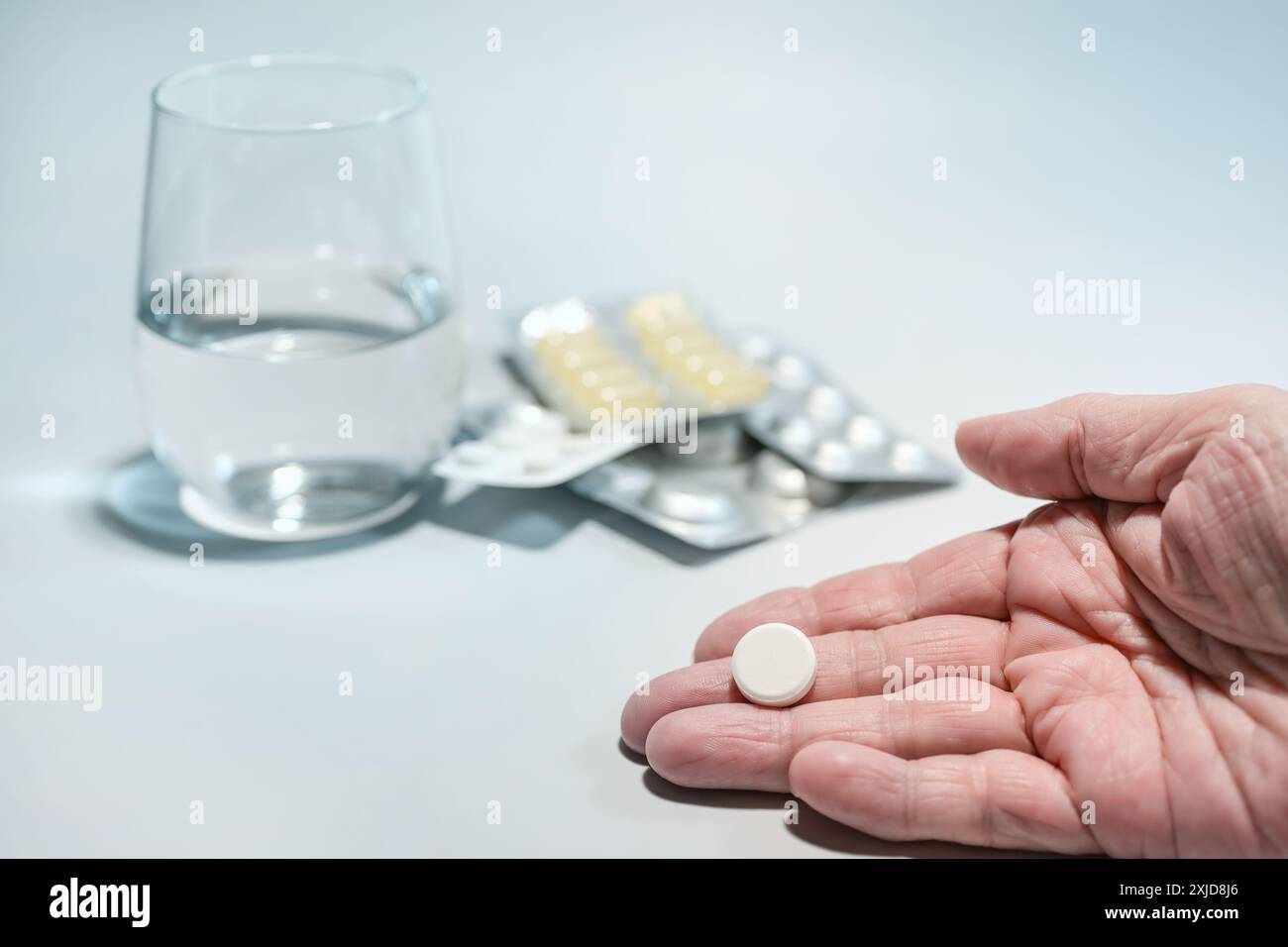 Ältere Hand mit einer Pille, verschwommenen Medikamentenpaketen und einem Glas Wasser im hellen Hintergrund, Konzept für Medizin und Gesundheitskonzept, Copy sp Stockfoto