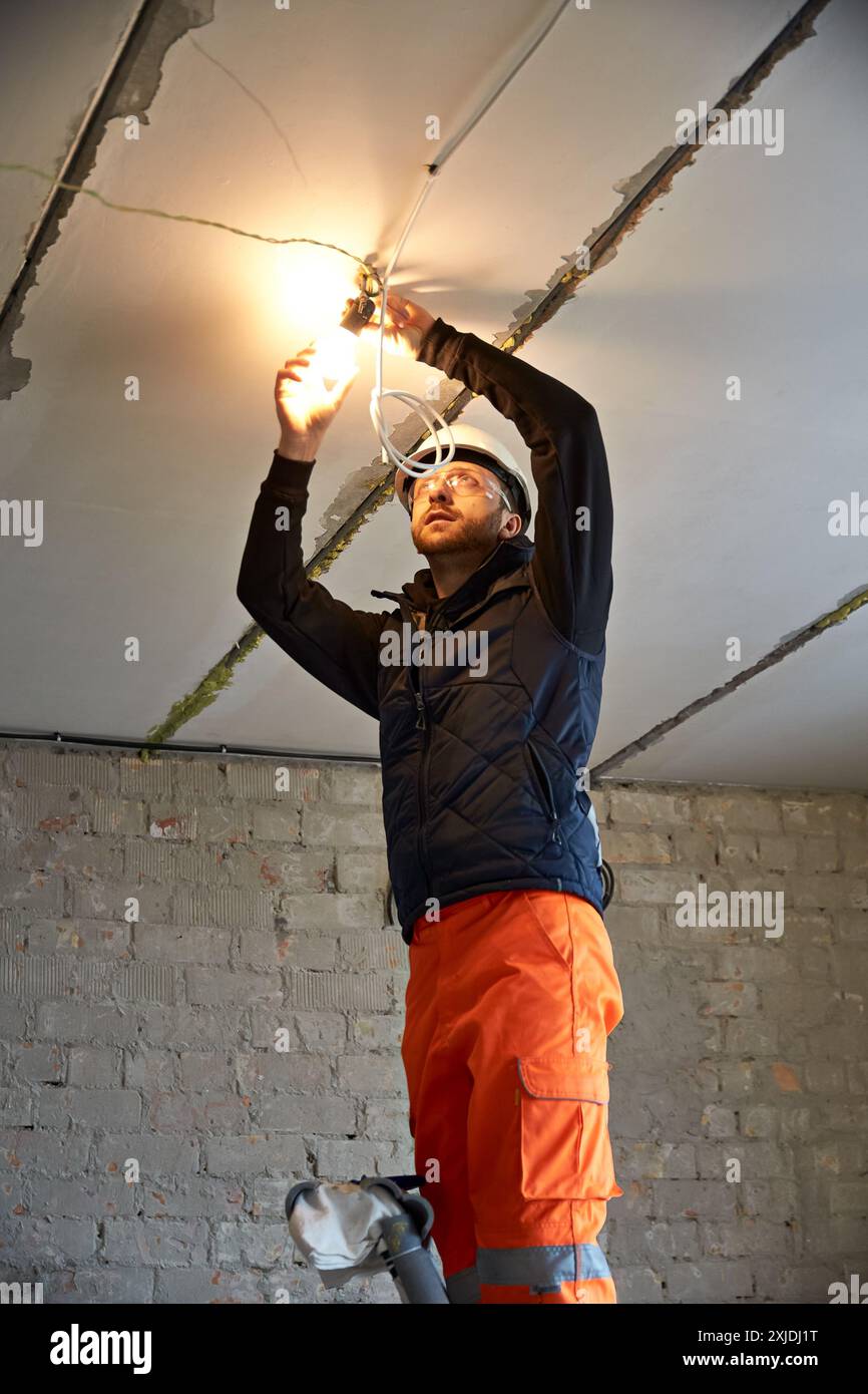 Elektriker mit weißem Schutzhelm und Schutzbrille, auf der Leiter stehend und konzentriert sich auf die Verkabelung einer Glühlampe an der Decke. Stockfoto