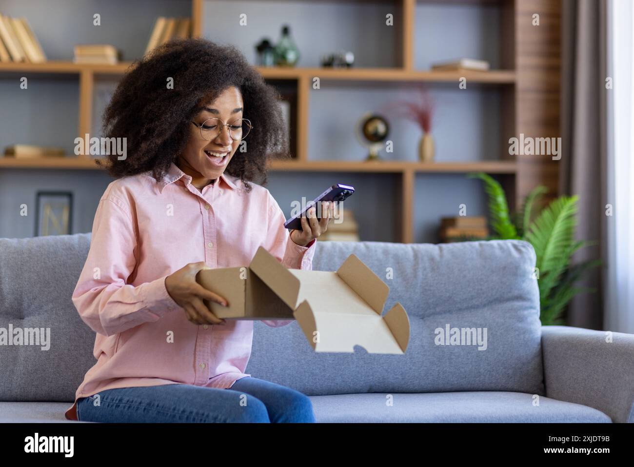 Glückliche junge Frau, die auf dem Sofa sitzt, während sie ihr Smartphone hält. Ich freue mich auf den Empfang von Bestellungen zu Hause. Konzept von Online-Shopping, Unboxing Experience und Kundenzufriedenheit. Stockfoto