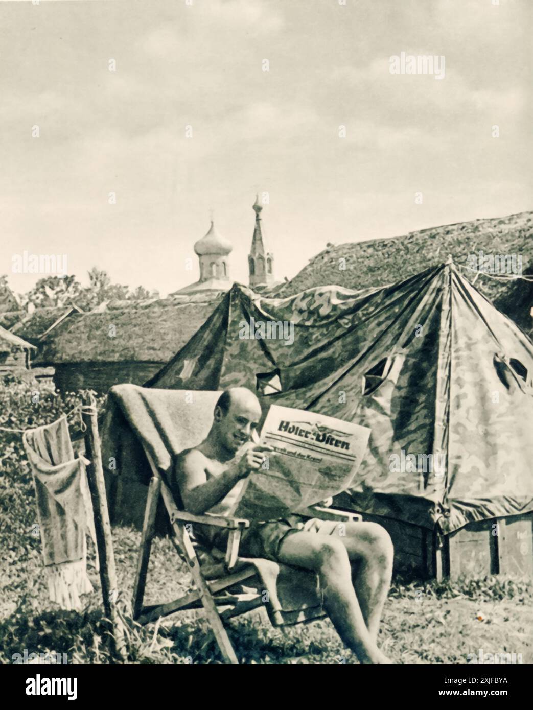 Ein Foto zeigt einen deutschen Soldaten, der in der Sonne sitzt und eine Zeitung im besetzten sowjetischen Territorium liest. Dieses Bild wurde 1941 während der Operation Barbarossa aufgenommen und zeigt einen kurzen Moment der Entspannung inmitten des unerbittlichen Vormarsches gegen die sowjetischen Truppen an der Ostfront. Stockfoto