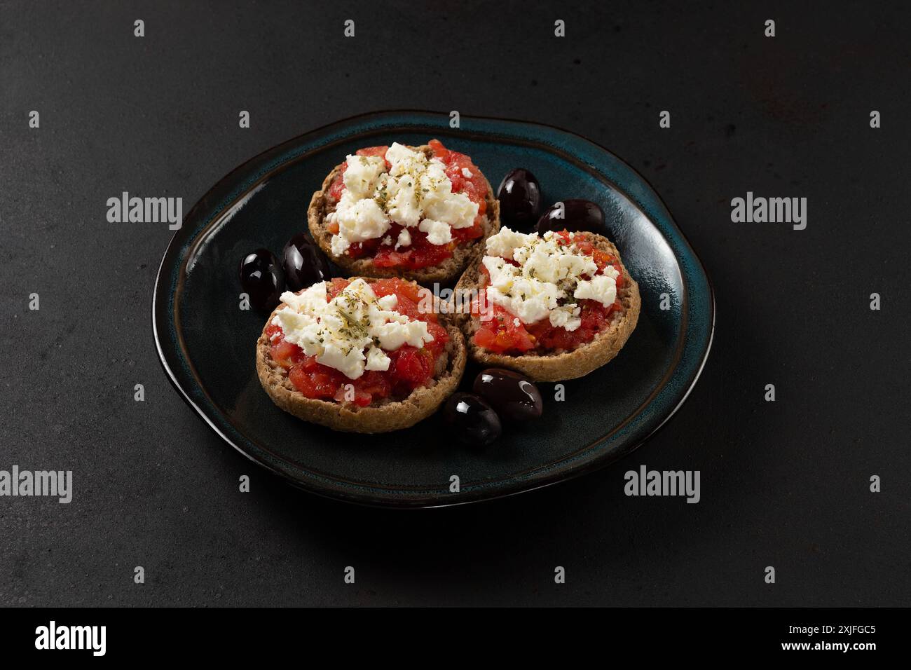 Nahansicht der kretischen Dakos auf dem Teller, bestehend aus gerstenrusk mit saftigen Tomaten, Fetakäse und Olivenöl auf dunklem Hintergrund. Stockfoto