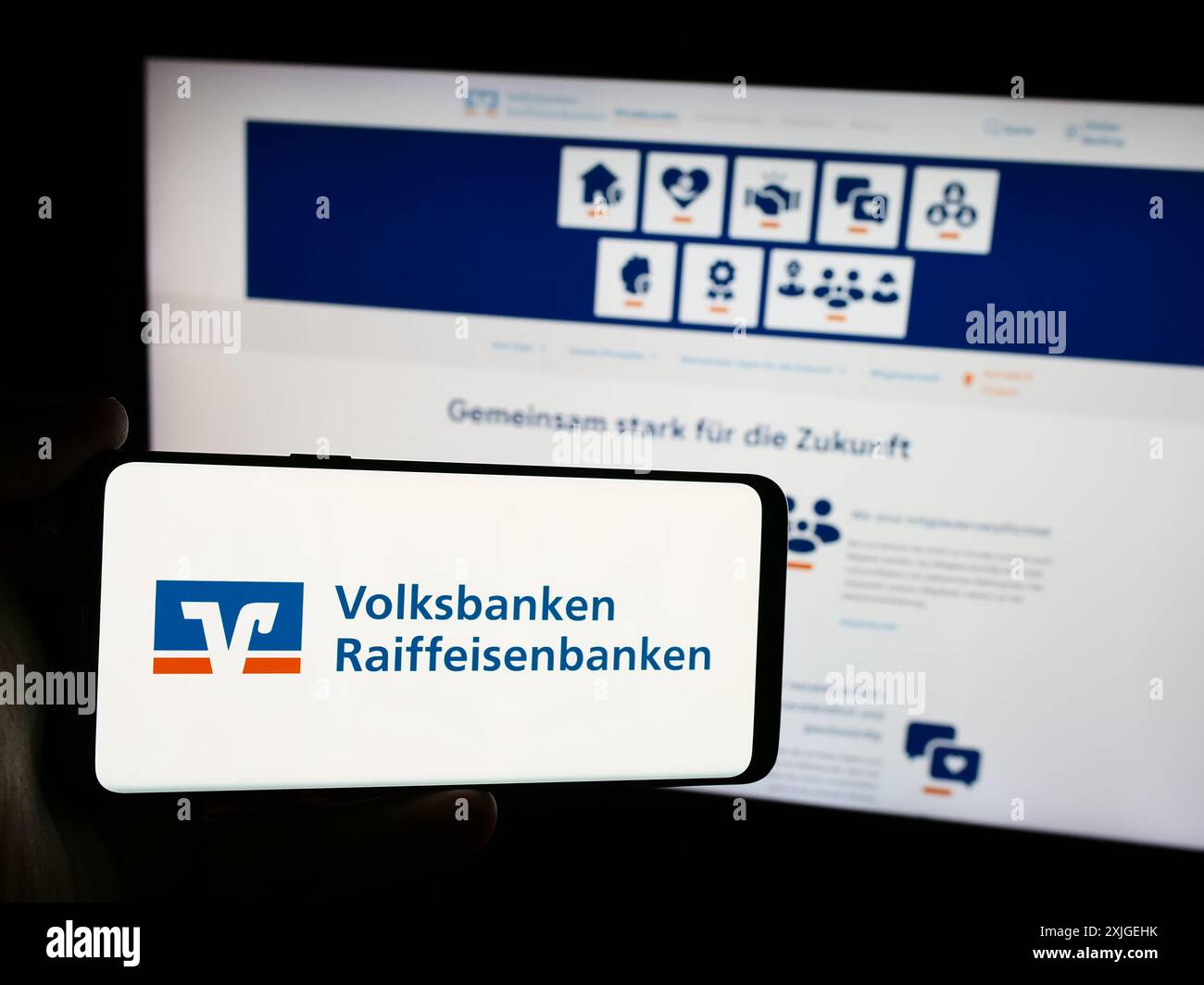 Person mit Mobiltelefon mit Logo des deutschen Banknetzes Volksbanken Raiffeisenbanken vor der Webseite. Schwerpunkt auf dem Display des Telefons. Stockfoto