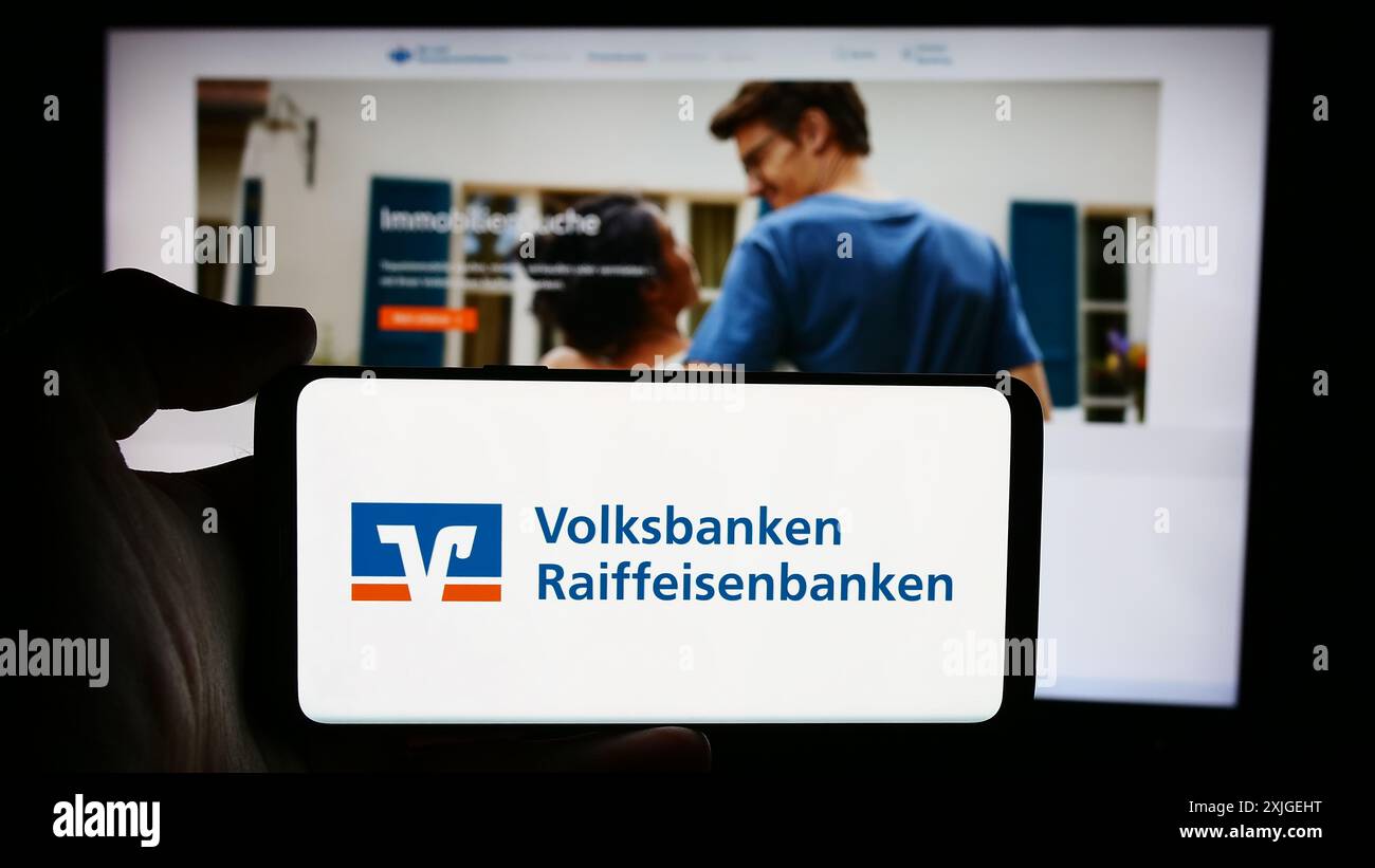 Person mit Handy mit Logo des deutschen Banknetzes Volksbanken Raiffeisenbanken vor der Business-Webseite. Schwerpunkt auf dem Display des Telefons. Stockfoto