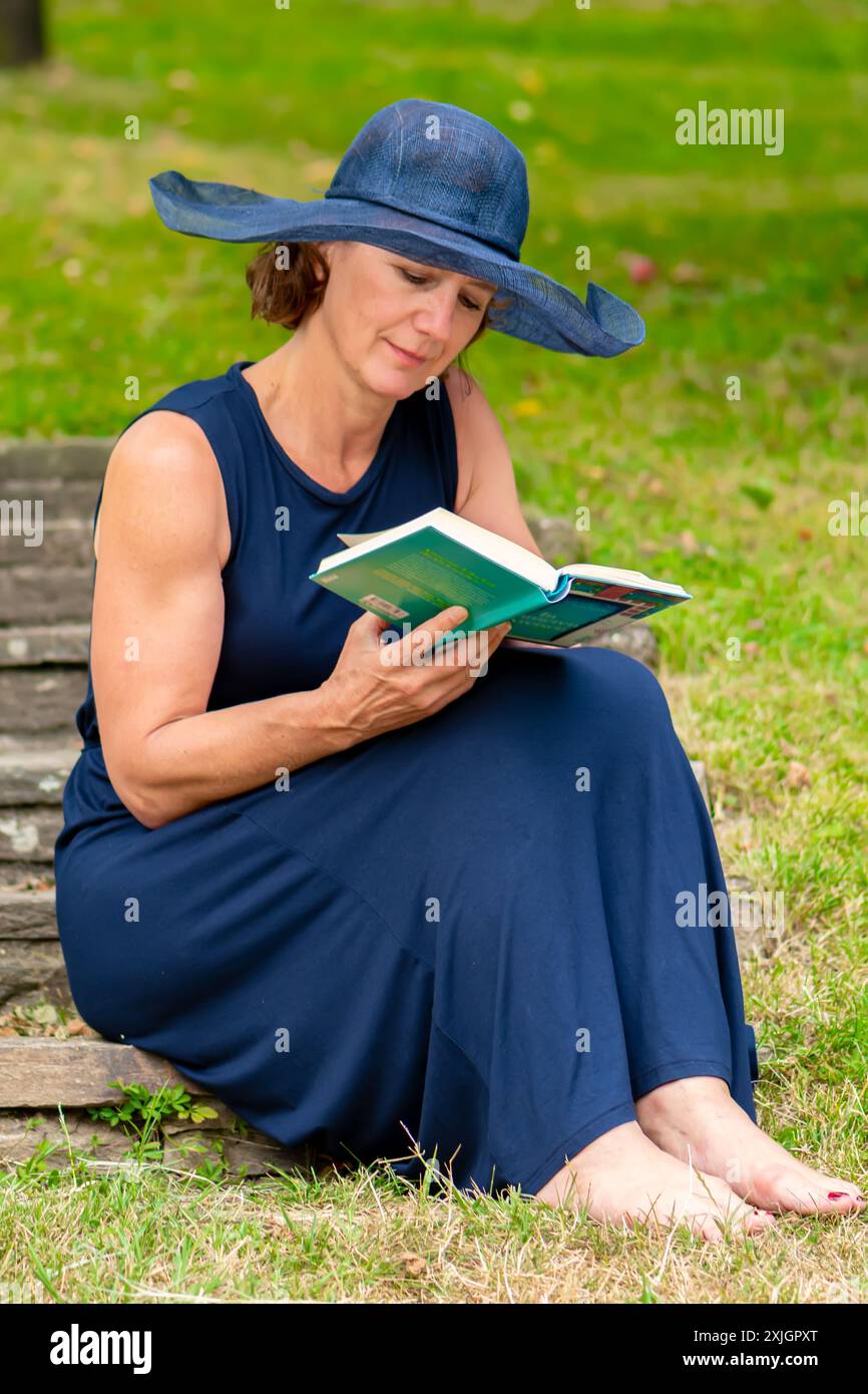 Eine 50-55-jährige Frau liest ein Buch auf einer Steintreppe in einem Sommergarten Stockfoto