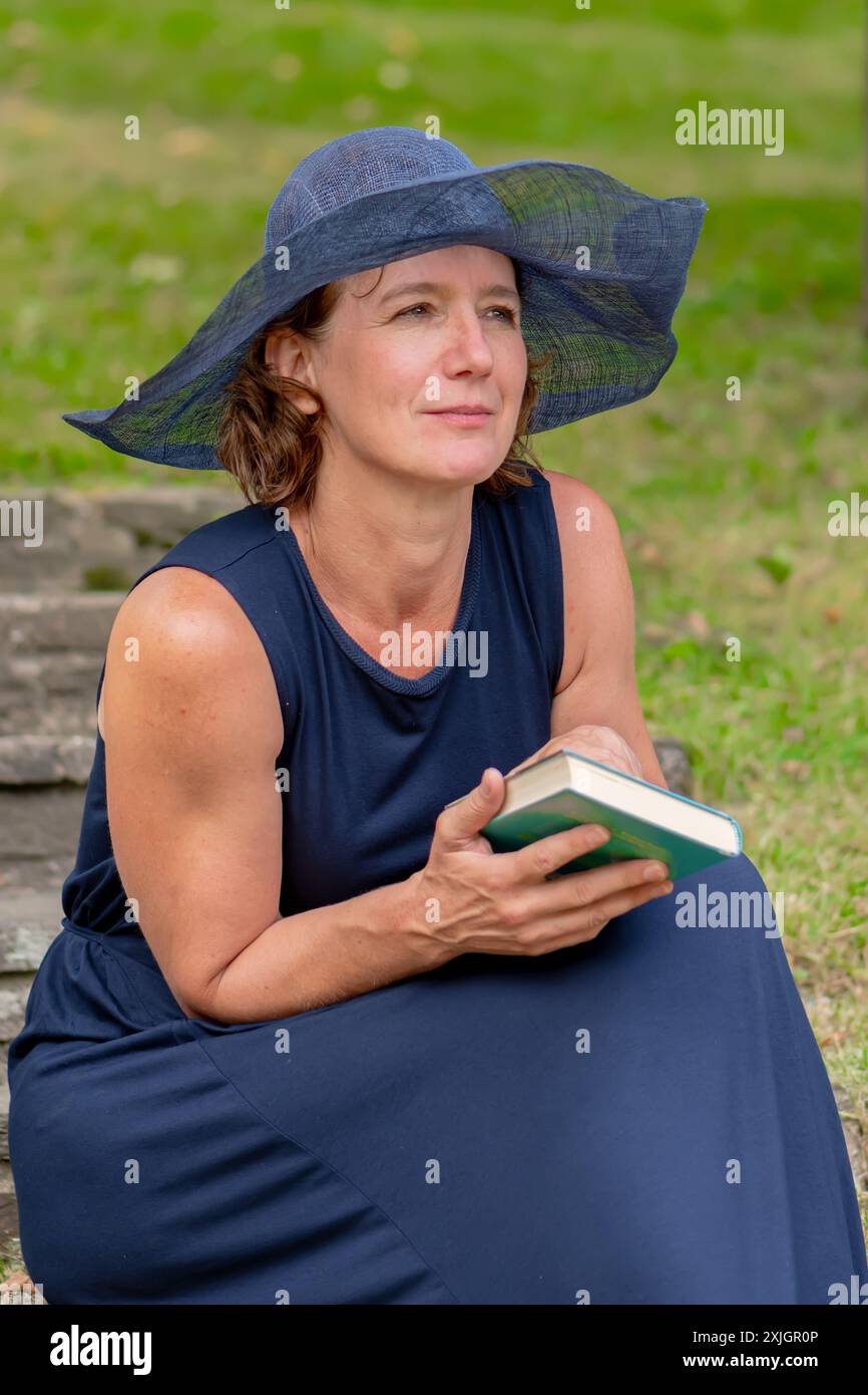 Eine Frau in blauem Kleid und breiter Krempe sitzt auf einer Steintreppe in einem grasbewachsenen Garten und hält ein Buch in der Hand. Stockfoto