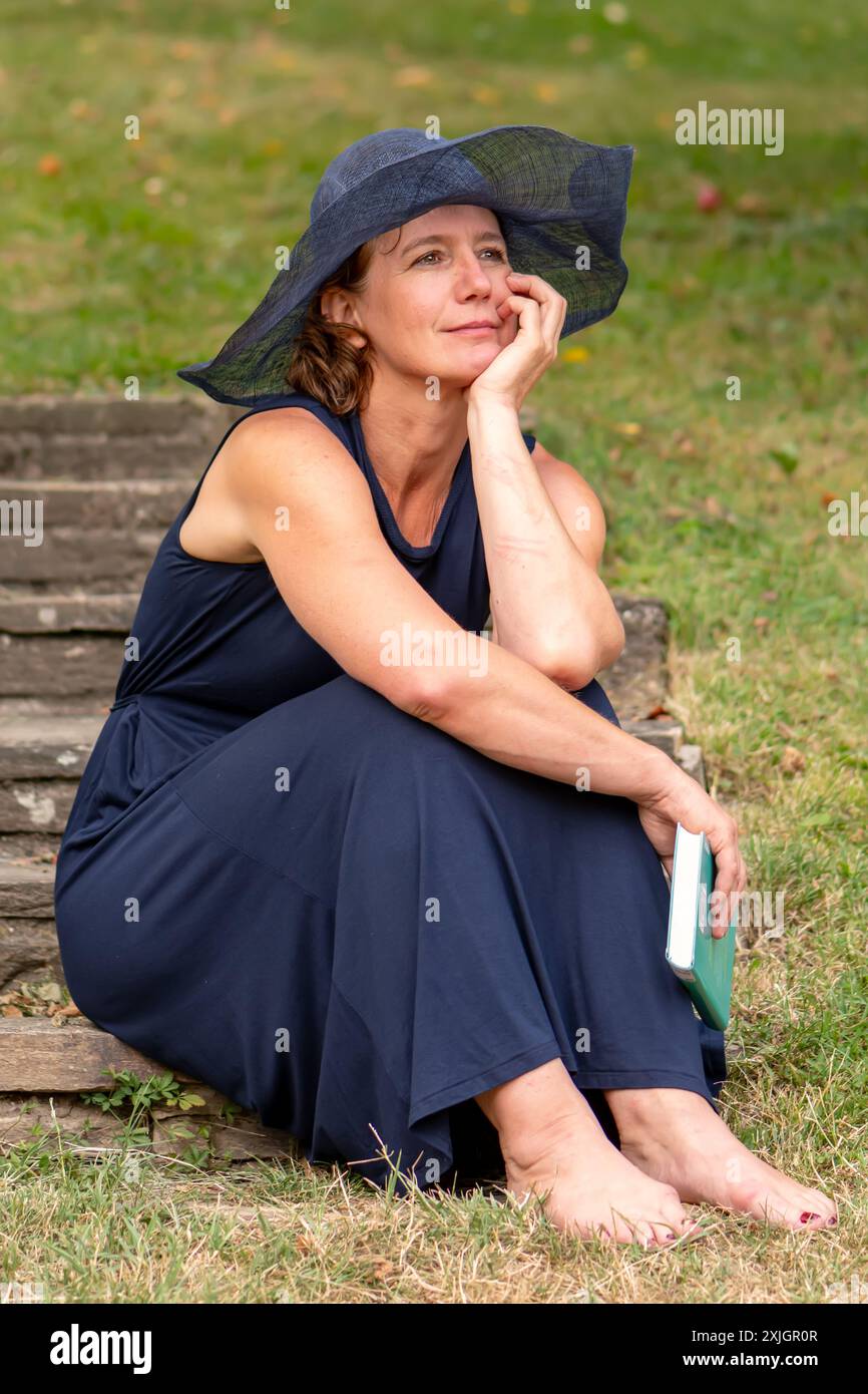 Porträt einer älteren Frau 50-55 Jahre alt mit blauem Damenhut und Kleid mit einem Buch in einem Sommergarten Stockfoto