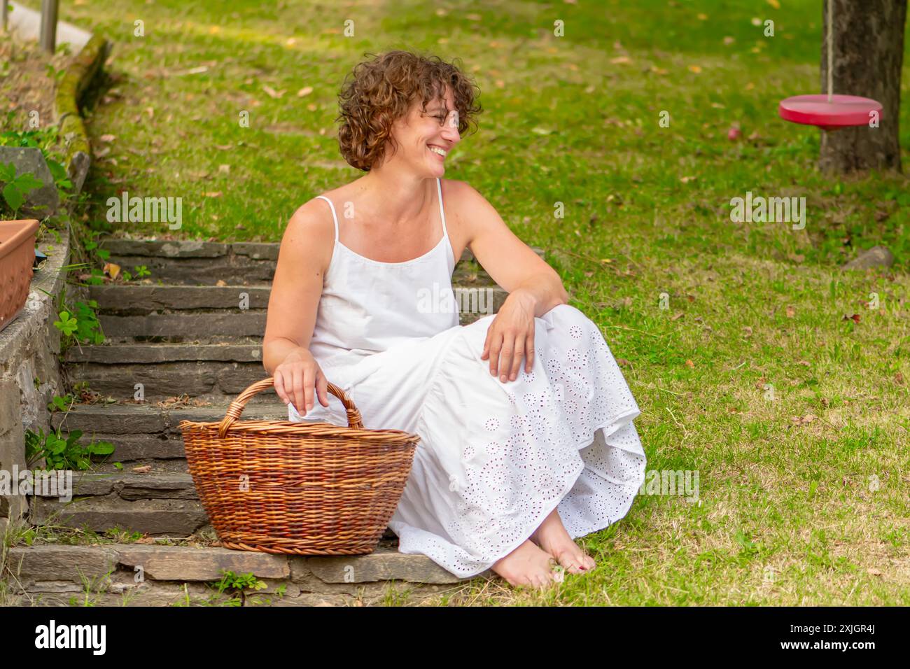 Eine Frau in einem weißen Kleid sitzt auf Steintreppen in einem grasbewachsenen Garten. Sie hat lockige Haare und ist barfuß. Ein Korbkorb steht auf der Treppe neben ihr. Stockfoto