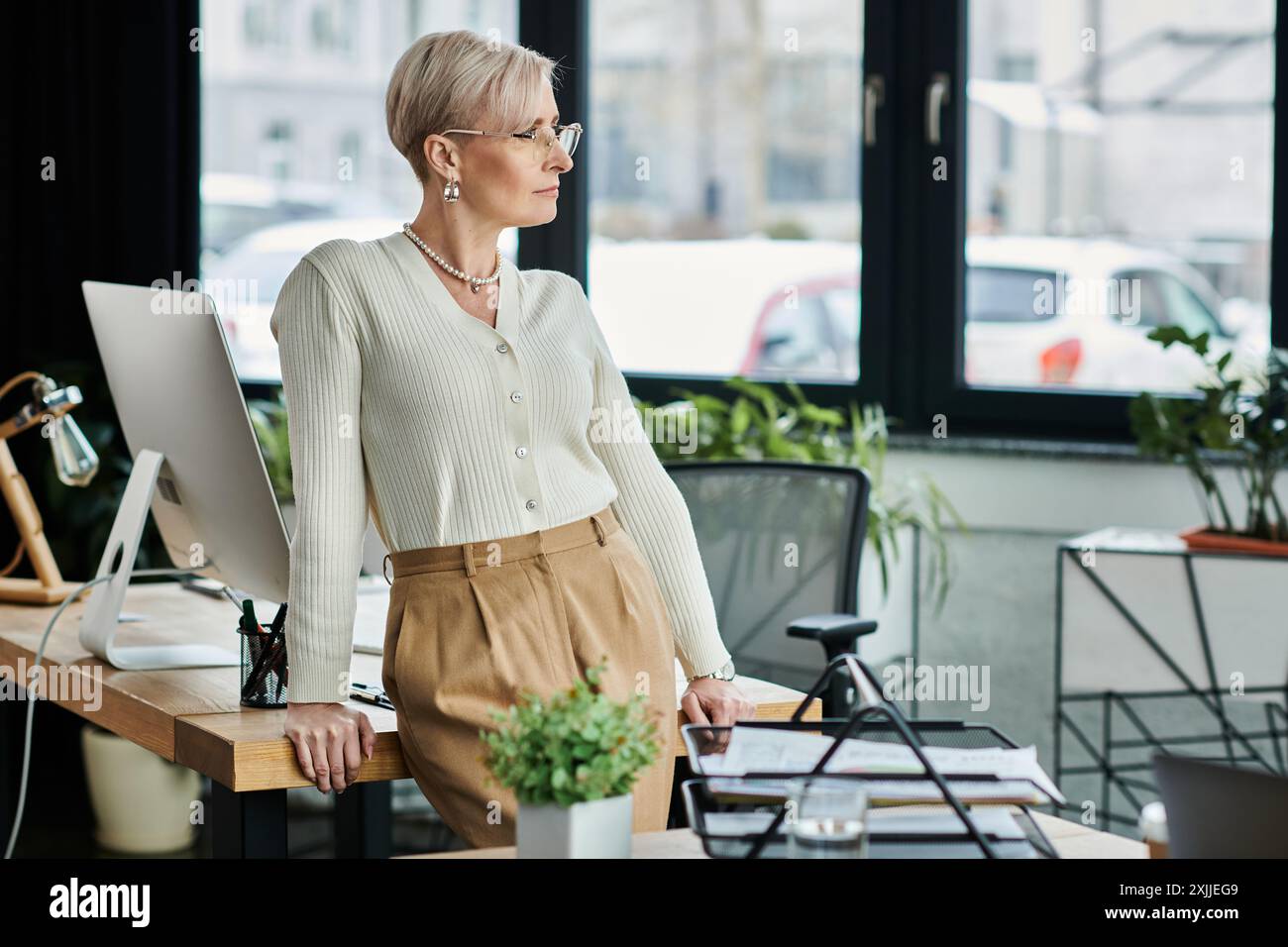 Eine Geschäftsfrau mittleren Alters mit kurzen Haaren steht auf einem eleganten Schreibtisch in einer modernen Büroumgebung. Stockfoto