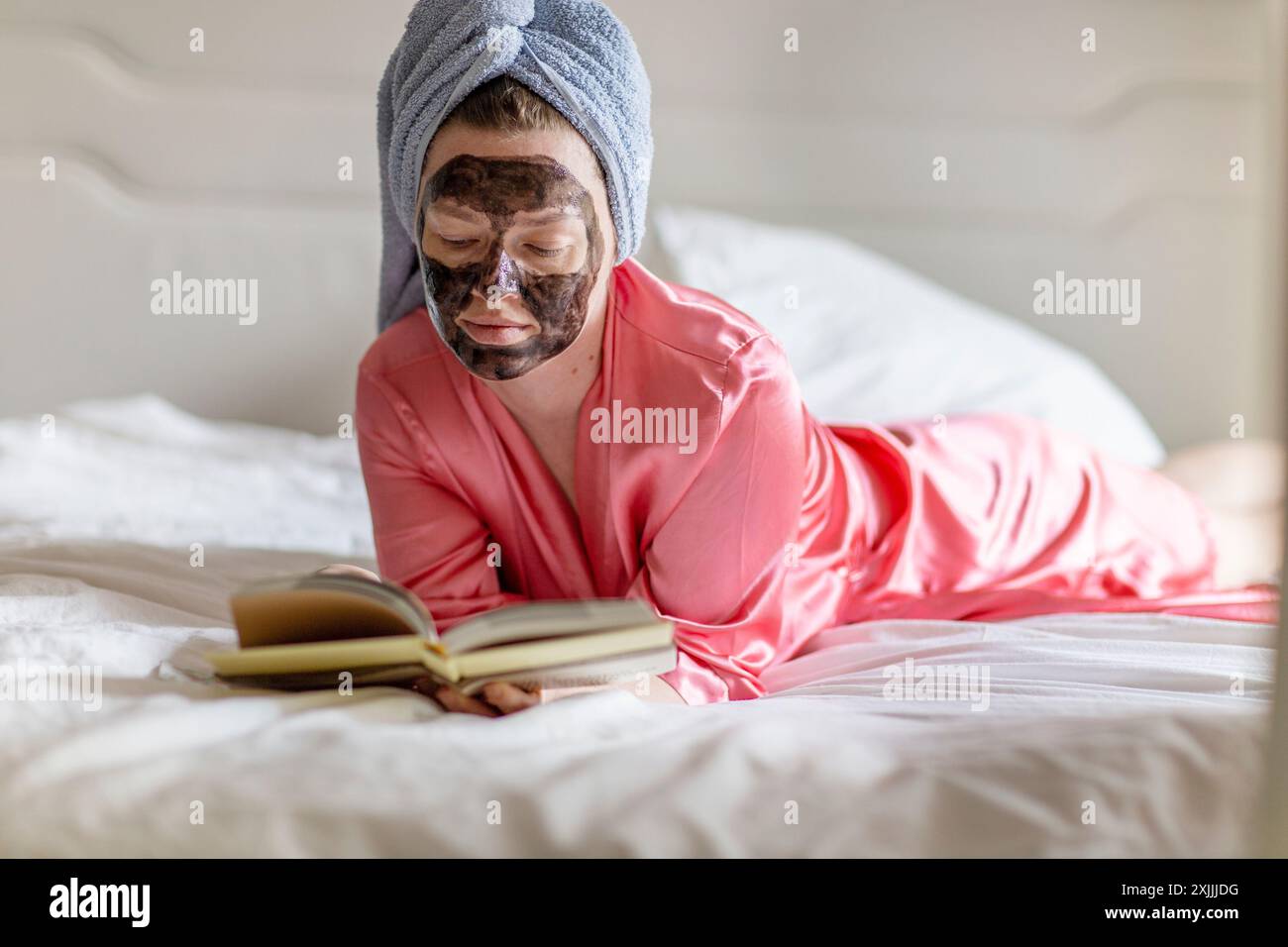Frau mit schwarzer Gesichtsmaske und Handtuch auf dem Kopf liest Buch auf dem Bett Stockfoto