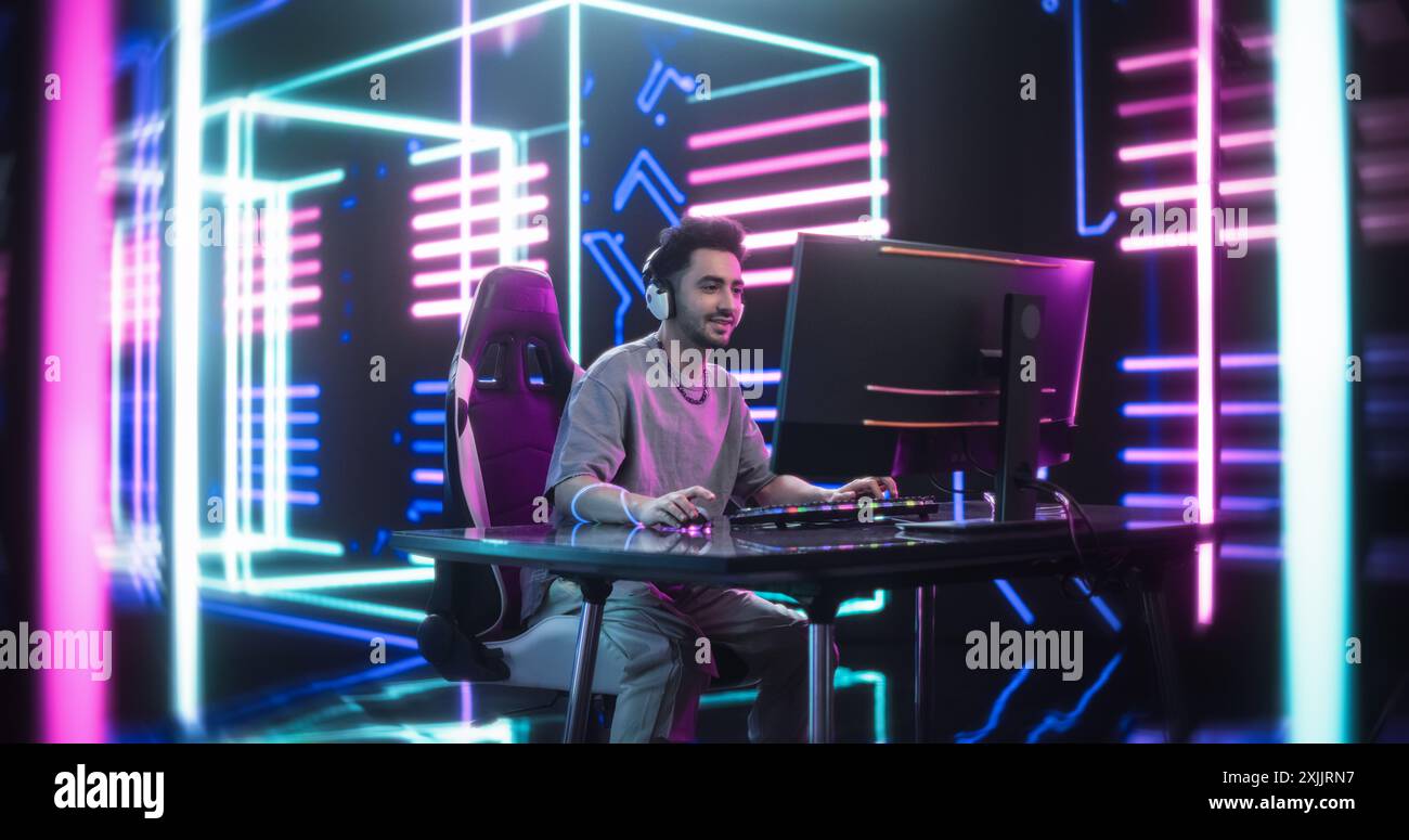 Junger Mann, der legere, bequeme Kleidung und eine Kette trägt, Videospiele auf einem Desktop-Computer spielt. Schöner Mann in Headphones streamt an ein Online-Publikum. Spiele in einem neonbeleuchteten Raum Stockfoto