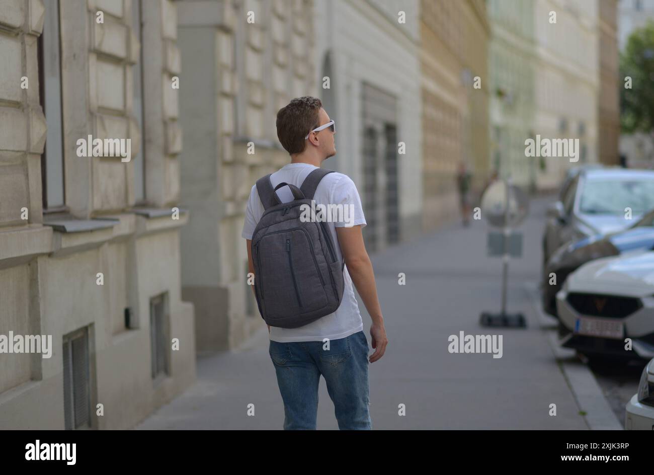 Ein Mann mit weißer Sonnenbrille und grauem Rucksack auf der Straße in Wien an einem heißen, sonnigen Sommertag. Tourismus- und Sightseeing-Konzept. Stockfoto