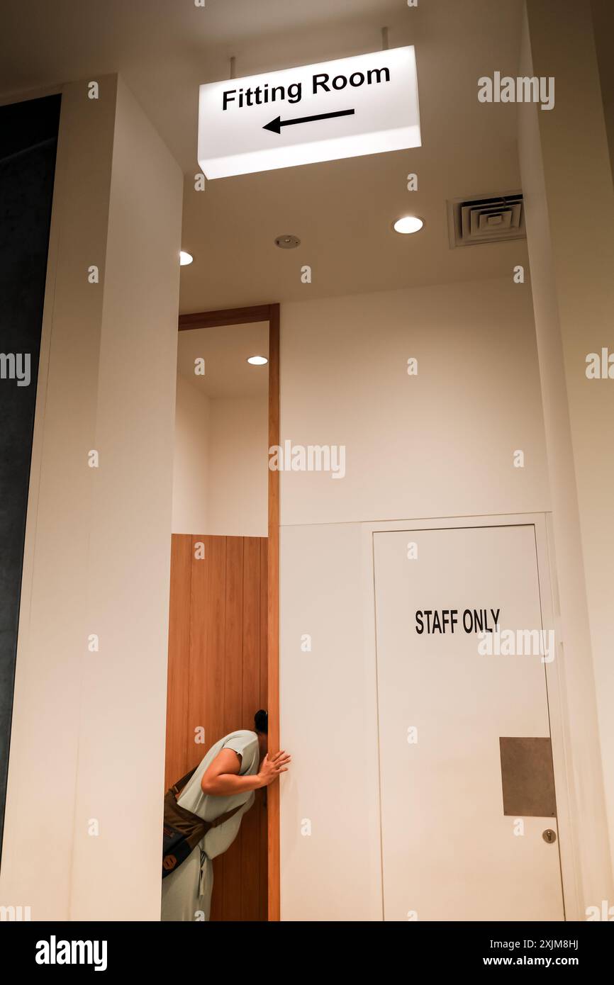 Eine erwachsene Frau, die in einen Umkleideraum eines Bekleidungsgeschäfts in einem Einkaufszentrum blickt, mit einem Leuchtkasten an der Decke Stockfoto