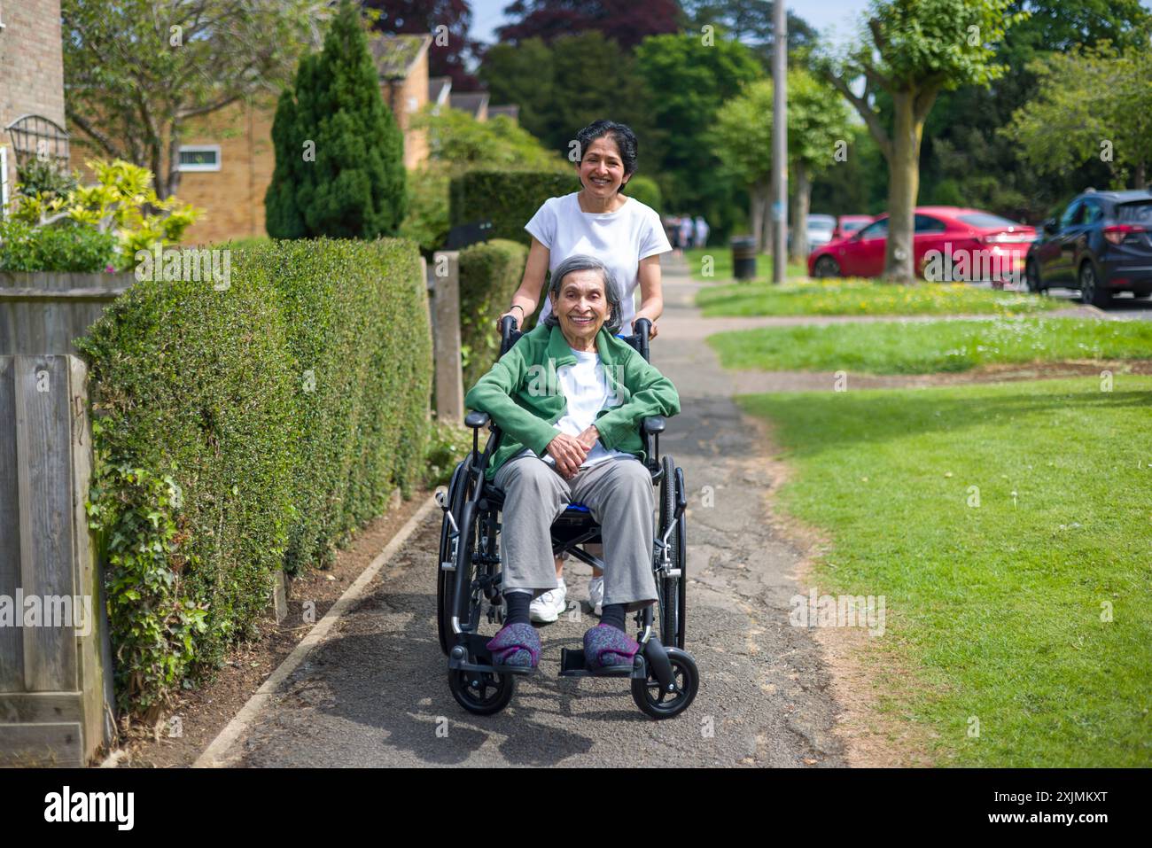 Alte indische Frau im Rollstuhl draußen auf einem Bürgersteig in einer Vorstadtstraße im Sommer, Großbritannien. Mutter und Tochter: Kann auch eine Betreuerin darstellen, die sich um die Ko kümmert Stockfoto