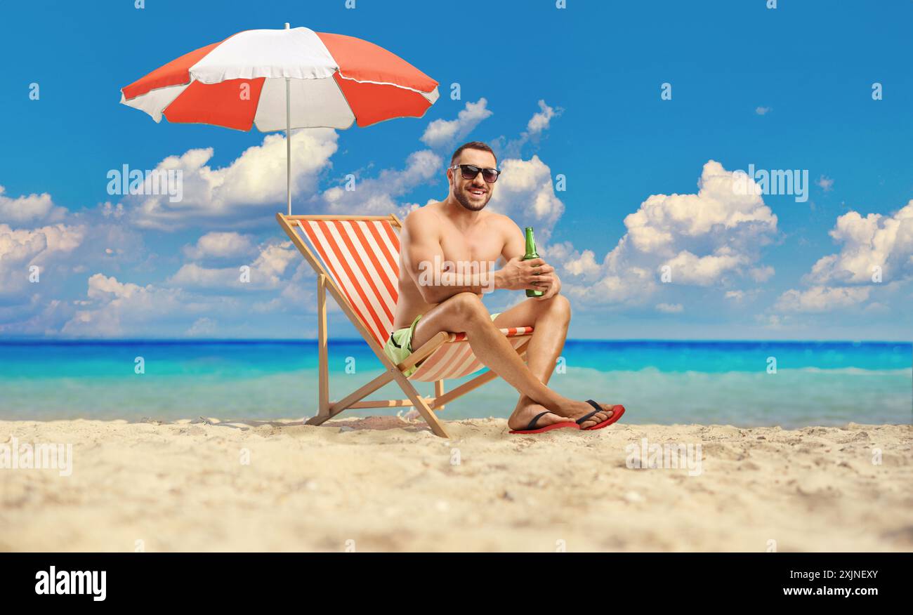 Ein Mann, der ein Bier am Sandstrand am Meer genießt, sitzt in einem Stuhl unter einem Sonnenschirm Stockfoto