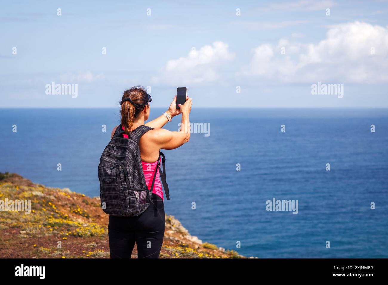 Frau fotografiert das Meer auf ihrem Smartphone, nachdem sie erfolgreich auf die Klippe der Algarve geklettert ist. Alleinreisen aktiver Sommerurlaub Portugal Stockfoto