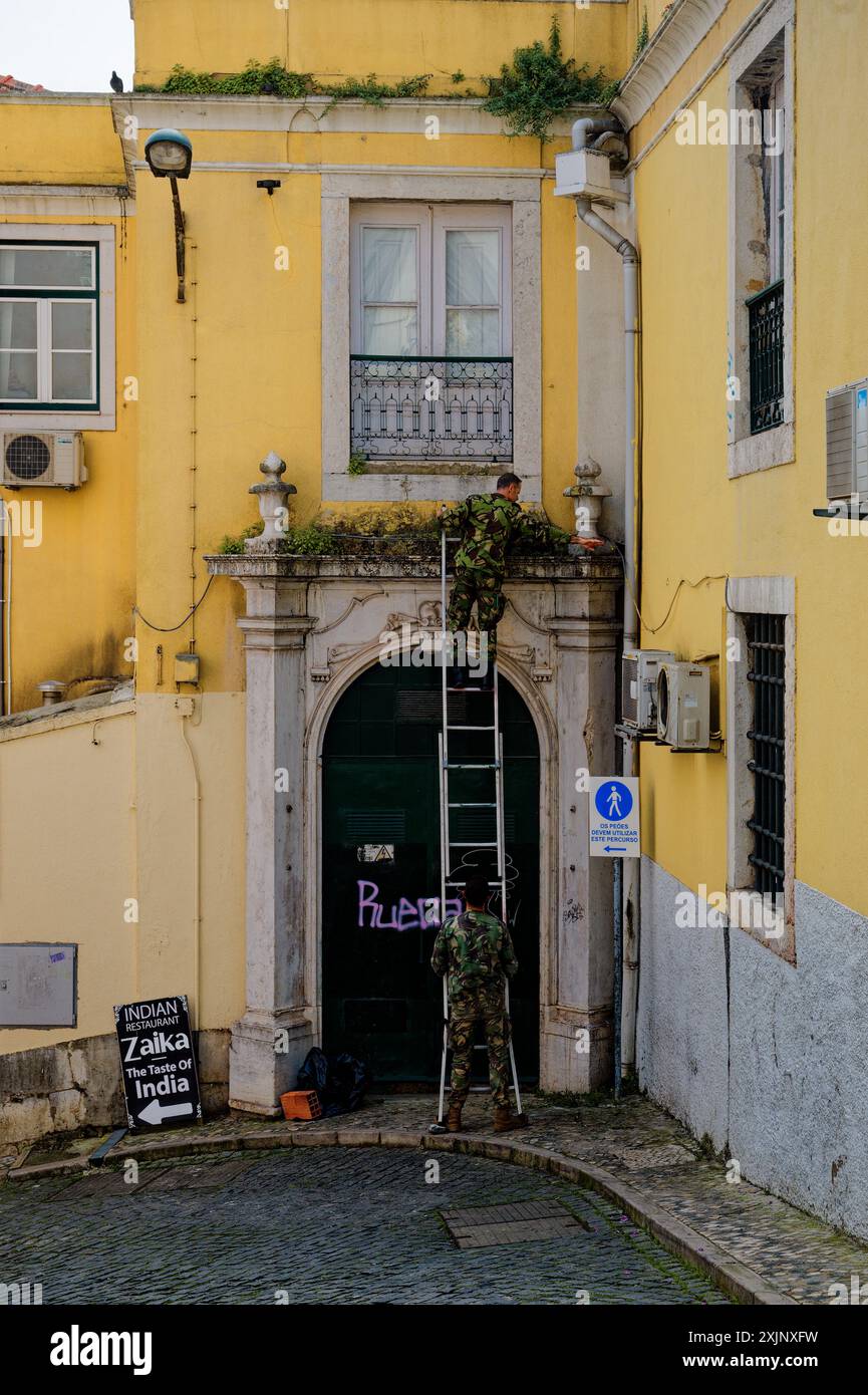 Zwei Soldaten in alfama, einer hält eine Leiter, während der andere wilde Pflanzen von einem überwachsenen alten Eingang entfernt Stockfoto