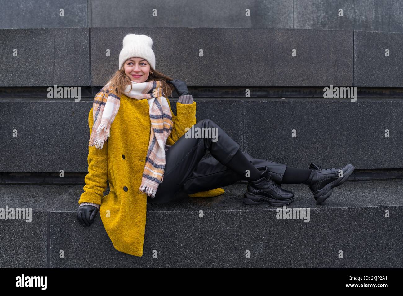 Eine stilvolle, elegante Frau in gelbem Mantel und weißem Hut sitzt auf einem Monument aus dunklem Granit. Sehenswürdigkeiten in Dresden, Deutschland Stockfoto