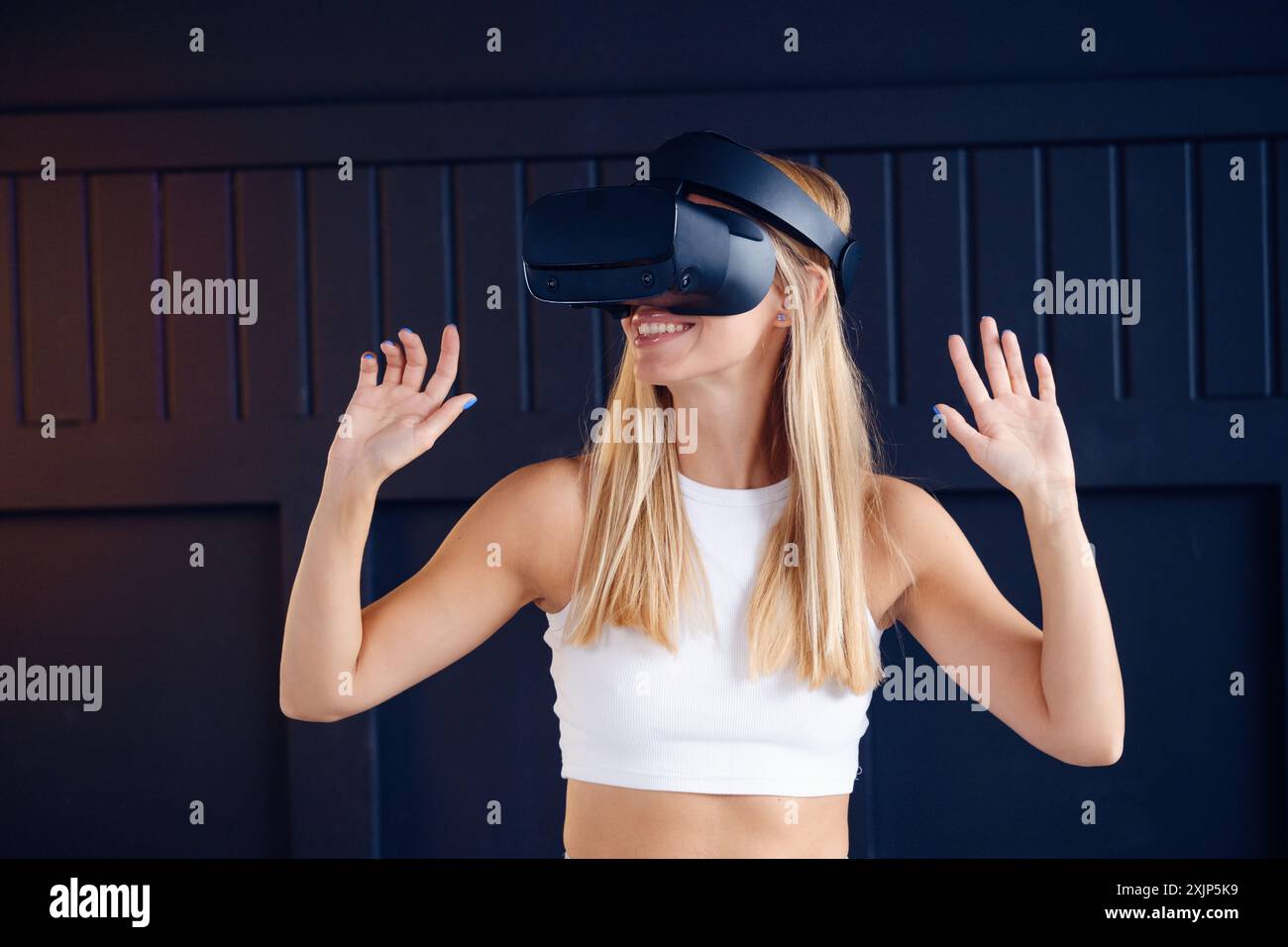 Die blonde Frau in weißem Tanktop lächelt mit dem VR-Headset vor der dunkelblauen Wand und interagiert mit der virtuellen Welt mit den Händen. Stockfoto