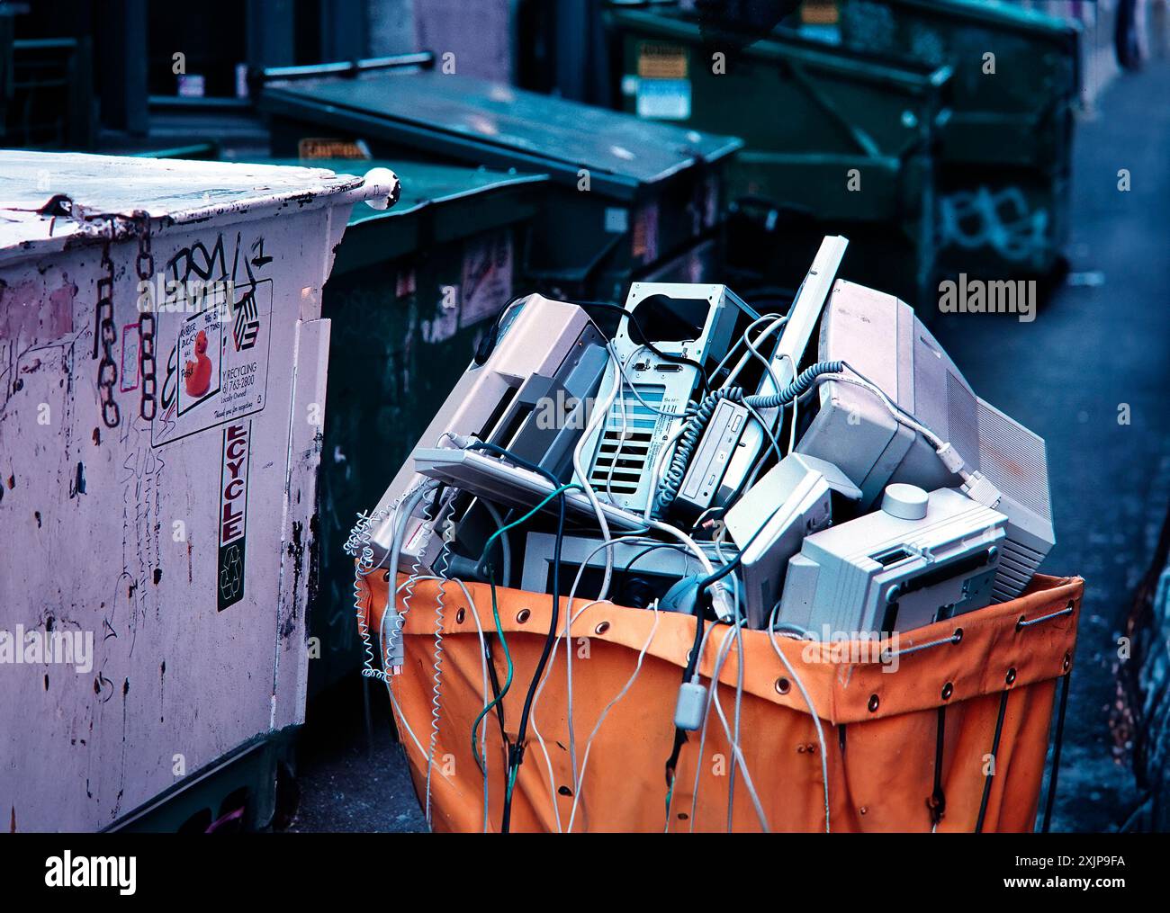 Ein gelber Papierkorb ist überfüllt mit weggeworfenen Computerkomponenten, einschließlich eines Monitors, eines Tower, einer Tastatur und anderer sonstiger Teile. Die Stockfoto