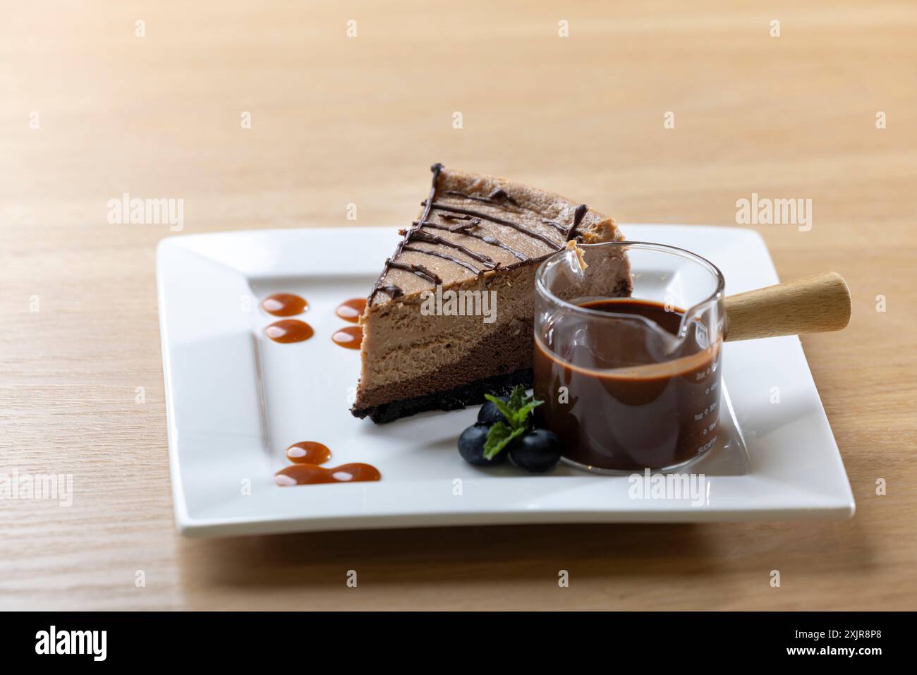 Dekadente Scheibe Schokoladen-Käsekuchen mit Schokoladensoße, serviert auf einem weißen quadratischen Teller. Begleitet von einer kleinen Tasse reichhaltigen Schokoladen-gan Stockfoto