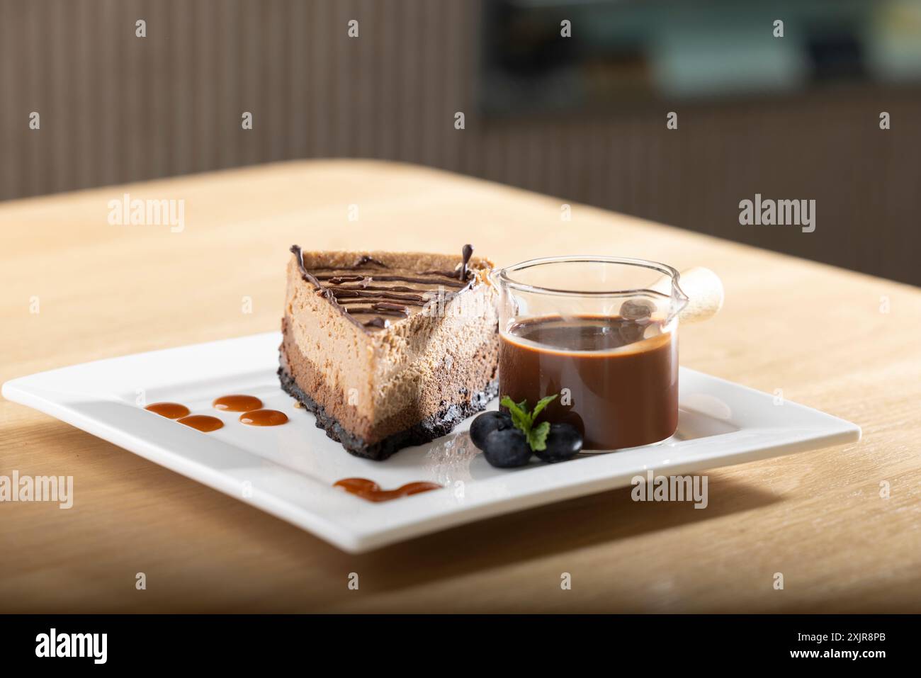 Dekadente Scheibe Schokoladen-Käsekuchen mit Schokoladensoße, serviert auf einem weißen quadratischen Teller. Begleitet von einer kleinen Tasse reichhaltigen Schokoladen-gan Stockfoto