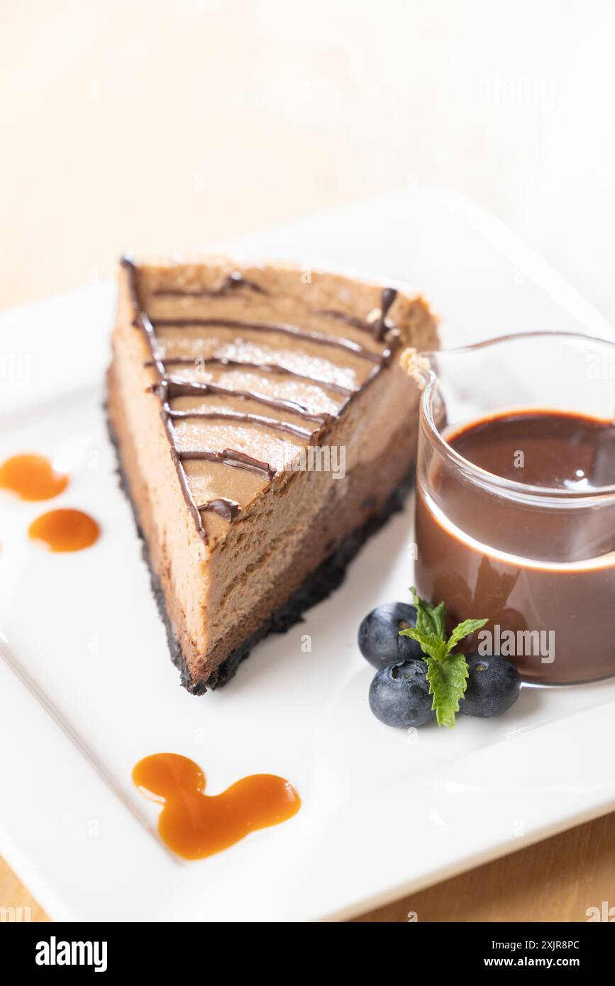 Dekadente Scheibe Schokoladen-Käsekuchen mit Schokoladensoße, serviert auf einem weißen quadratischen Teller. Begleitet von einer kleinen Tasse reichhaltigen Schokoladen-gan Stockfoto