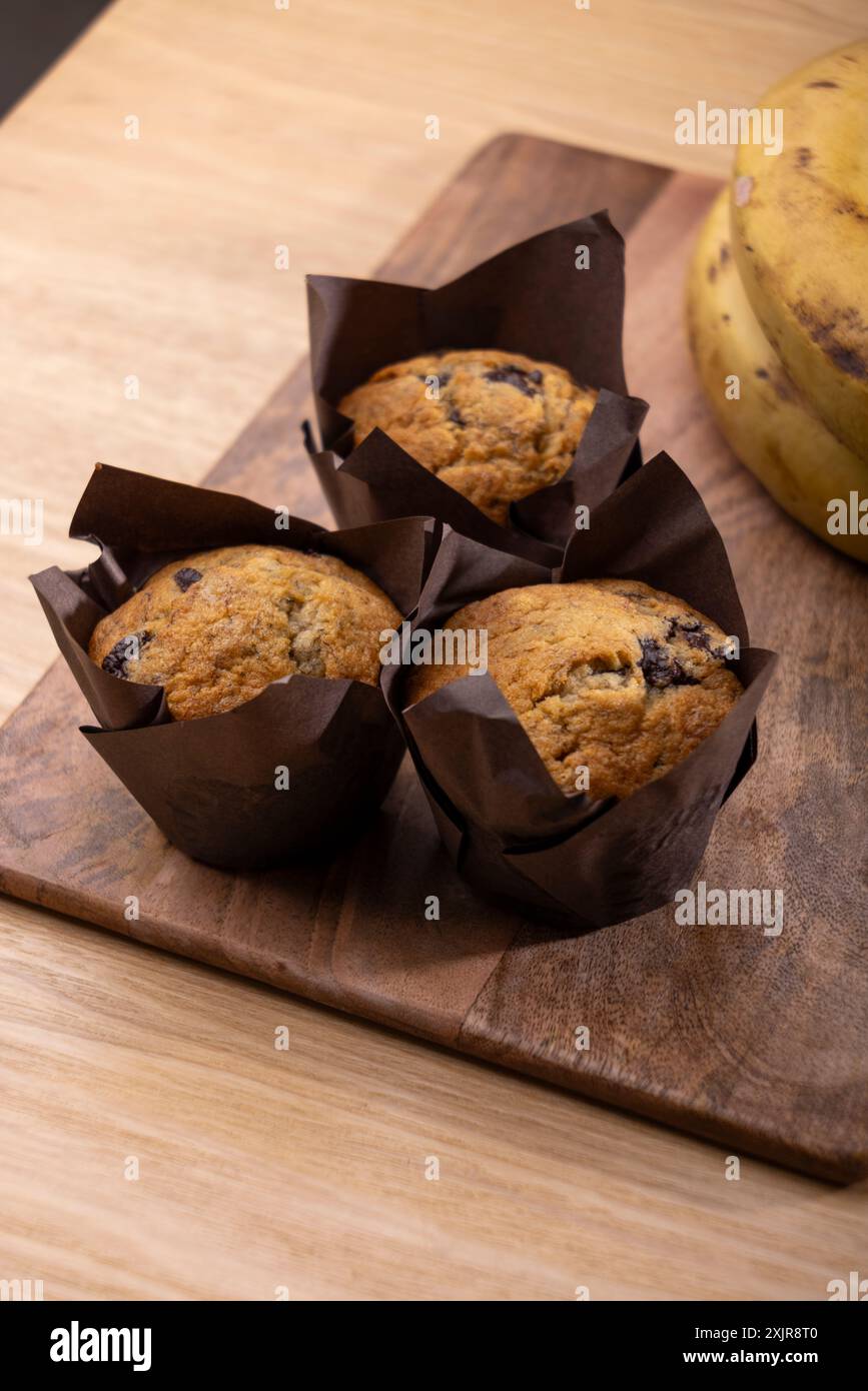 Frisch gebackene Bananenschokolade-Chips-Muffins in braunem Pergamentpapier, auf einem Holzschneidebrett neben einem Haufen reifer Bananen. Stockfoto
