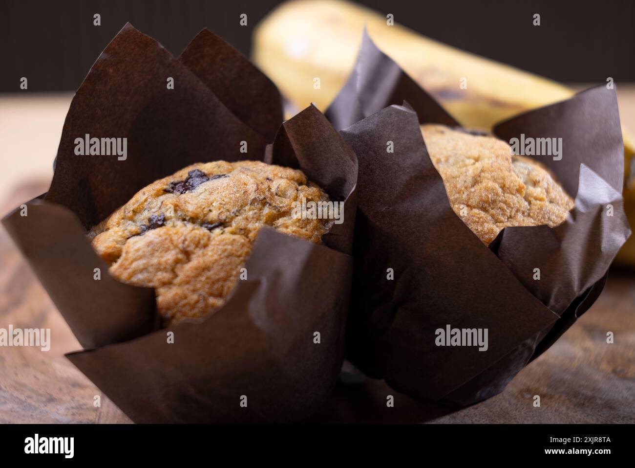 Frisch gebackene Bananenschokolade-Chips-Muffins in braunem Pergamentpapier, auf einem Holzschneidebrett neben einem Haufen reifer Bananen. Stockfoto