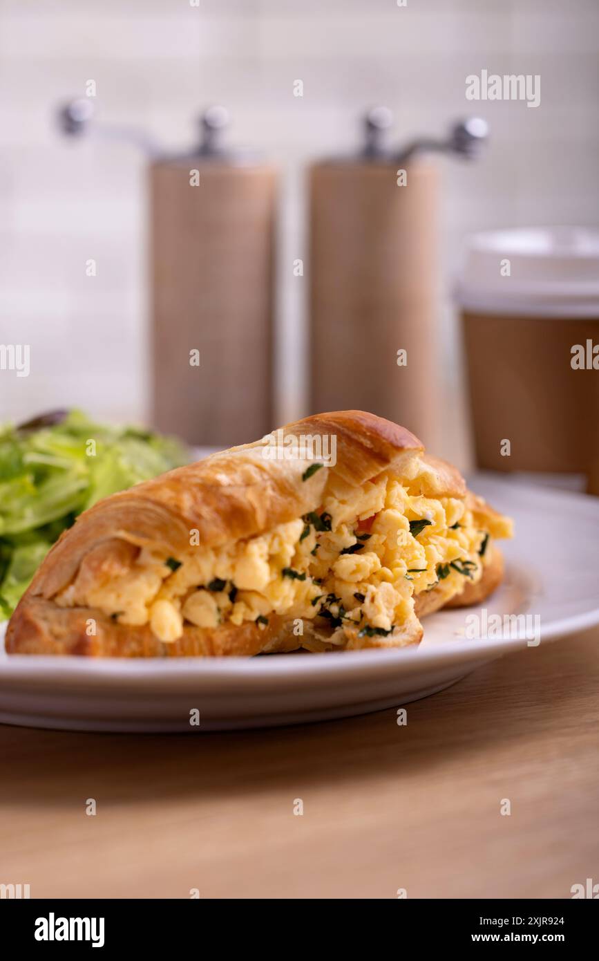 Ein Frühstücksangebot mit einem Croissant mit Rührei und Kräutern, serviert mit einem Salat auf einem weißen Teller. Stockfoto